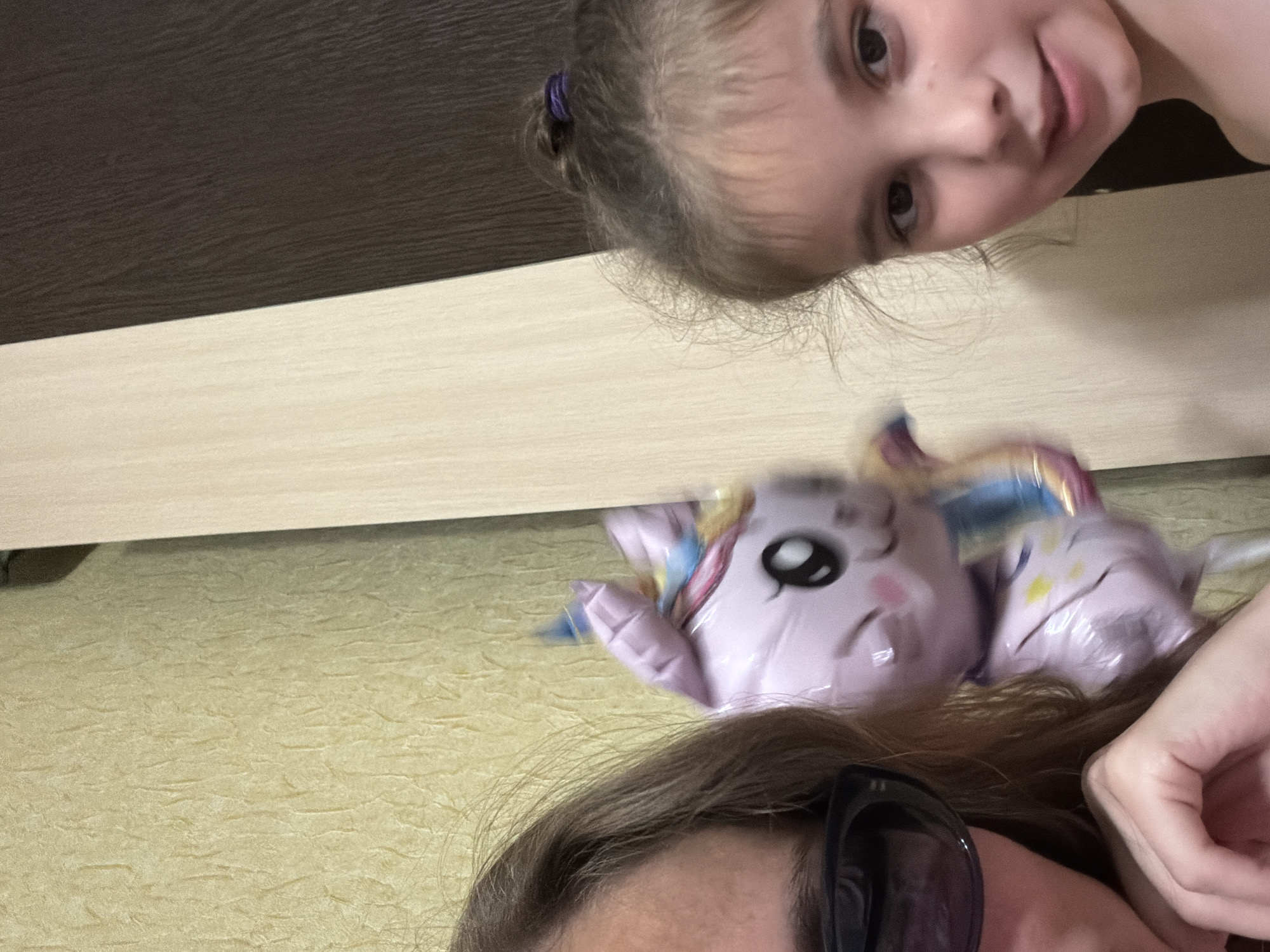 The image is a selfie taken indoors, featuring two individuals. On the left, a person partially visible wears sunglasses and has long brown hair. In the center, a pink unicorn balloon is blurred due to motion. On the right, a young girl with light brown hair tied in a bun looks at the camera. The background consists of textured yellow wallpaper on the left and a two-toned vertical divider, with a light-colored section on the left and a dark brown section on the right. The scene does not provide any specific details to indicate its location within the city of Tambov, Russia.