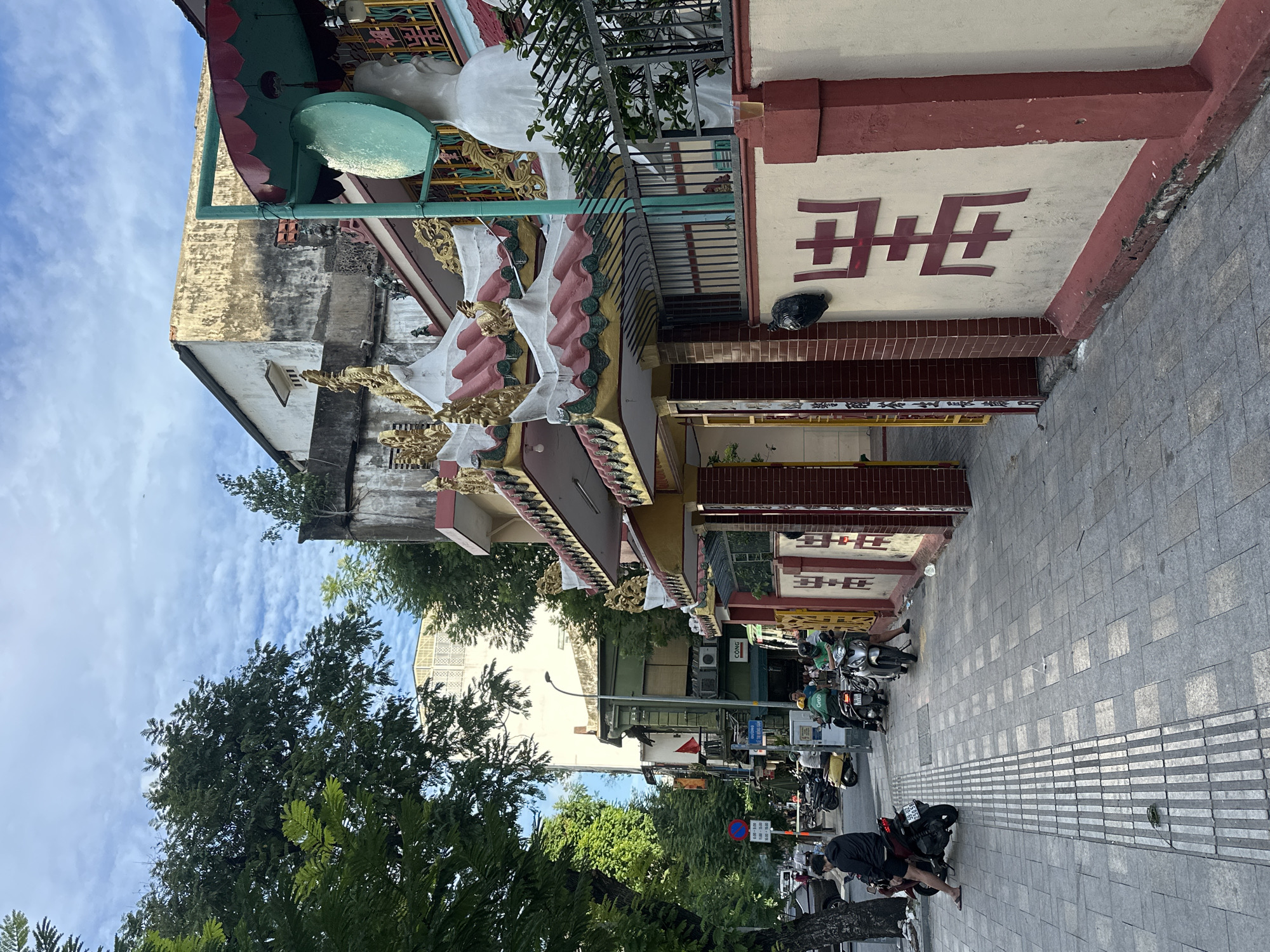 The image depicts a street scene in Thủ Đức, Vietnam, likely in the morning or afternoon given the bright sky and shadows. The weather appears to be clear with scattered clouds.
The foreground features a paved sidewalk with a textured tactile paving strip. Several motorcycles are parked along the street. Two people are visible in the lower right corner, one sitting on a motorcycle and another crouching nearby. Further down the street, more motorcycles and a few people can be seen, suggesting a bustling urban environment.
The background is dominated by a building with ornate, traditional architecture. It features a decorative facade with red and white paint, intricate gold trim, and what appear to be stylized carvings or figures. A large, circular green awning is visible on the left side of the building. The building has a weathered appearance, suggesting it is an older structure. A prominent red symbol is affixed to the building's exterior wall, with Chinese characters also visible on the building's ornamentation. Text on a sign on the building reads "CONC". A red flag is also visible further down the street. The overall impression is of a vibrant cultural and commercial area in Vietnam.