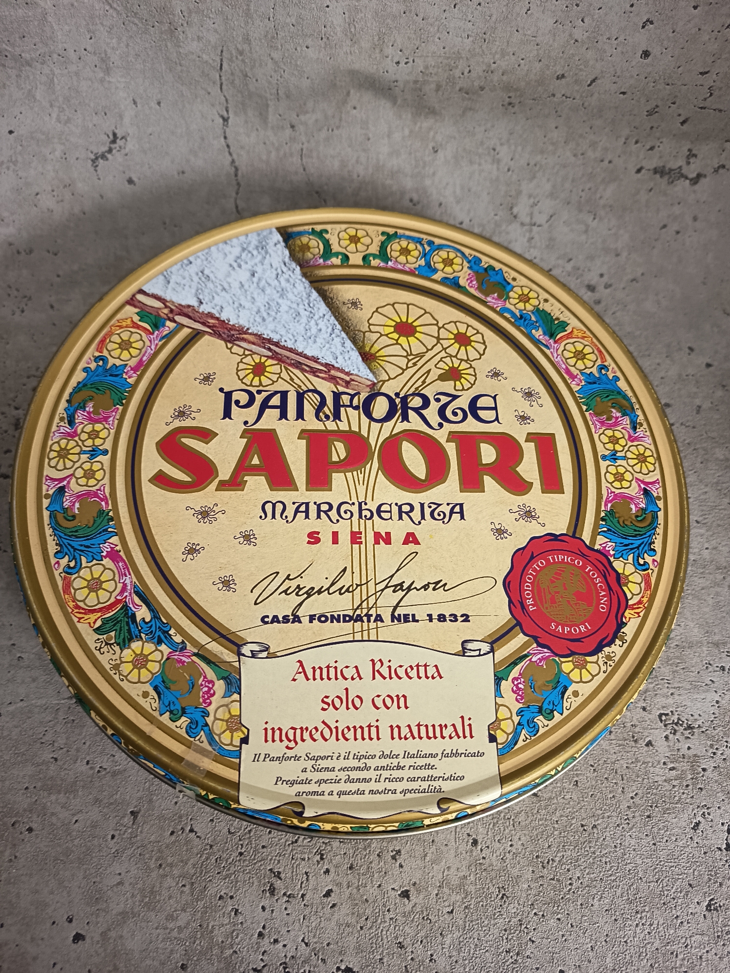 The image shows a round, metal tin of Panforte Sapori, a traditional Italian sweet. The tin's design features a gold background with floral patterns and the brand name prominently displayed in red letters. There is a wedge cut out of the tin, exposing a powdery, white substance. The text on the tin indicates that it is made with natural ingredients and is a specialty from Siena. The background appears to be a gray, textured surface. The image does not show the location of Wangen bei Olten, Switzerland, nor does it contain any elements suggesting the location.