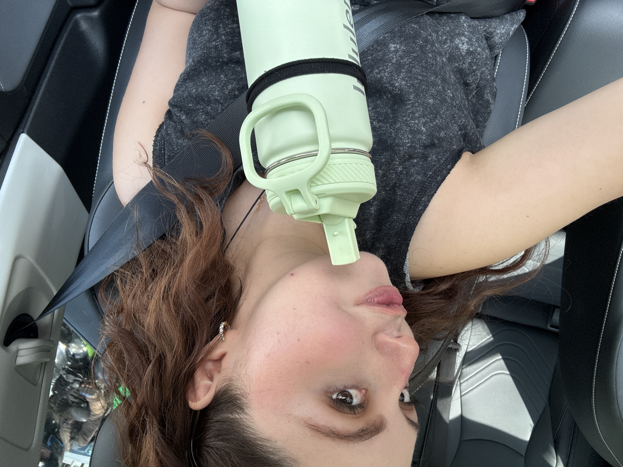 A woman with brown wavy hair is seated in a car, secured by a seatbelt, with a light green water bottle positioned above her face. The interior of the car is predominantly black and grey, with white stitching on the seats. The background visible through the window suggests an urban environment, although specific details are indistinct. The city cannot be determined based solely on the information in the image.