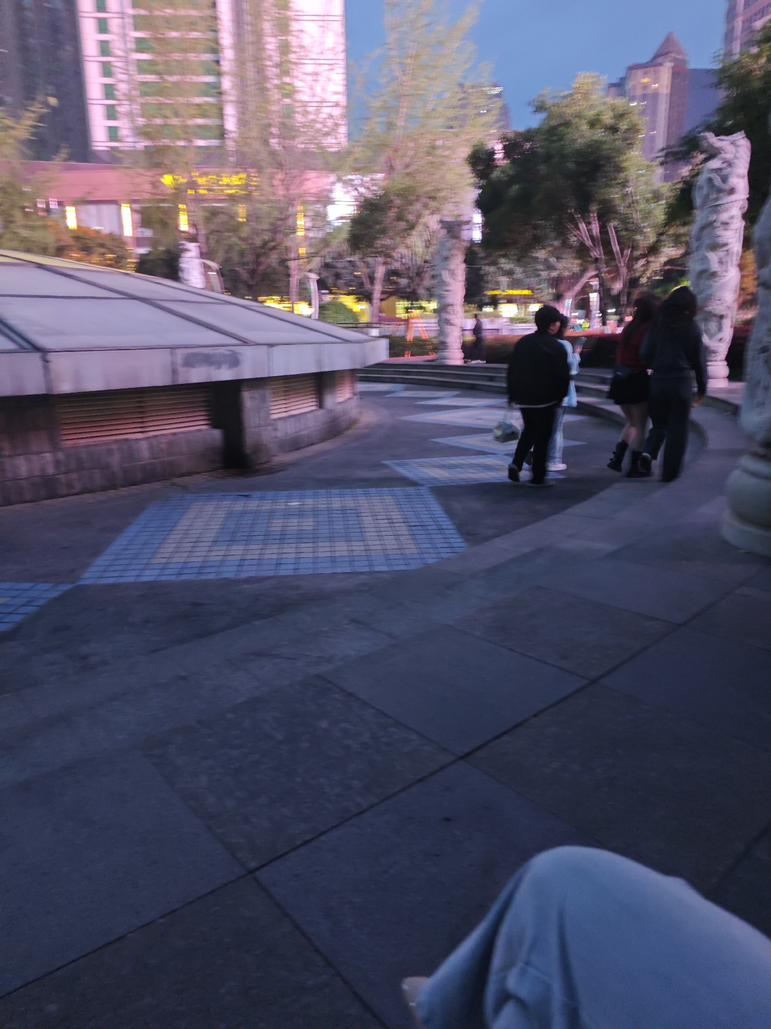 The image depicts an urban scene in Liangjiang, China, at twilight. Three people are seen walking on a paved area with blue and white checkered patterns towards the right side of the frame. To their left is a low building with a glass roof. In the background, there are trees and buildings, including high-rise structures illuminated with artificial lights. To the far right are carved stone columns. The bottom right corner of the image shows a person sitting with their knee visible. The overall environment appears to be a public square or park.