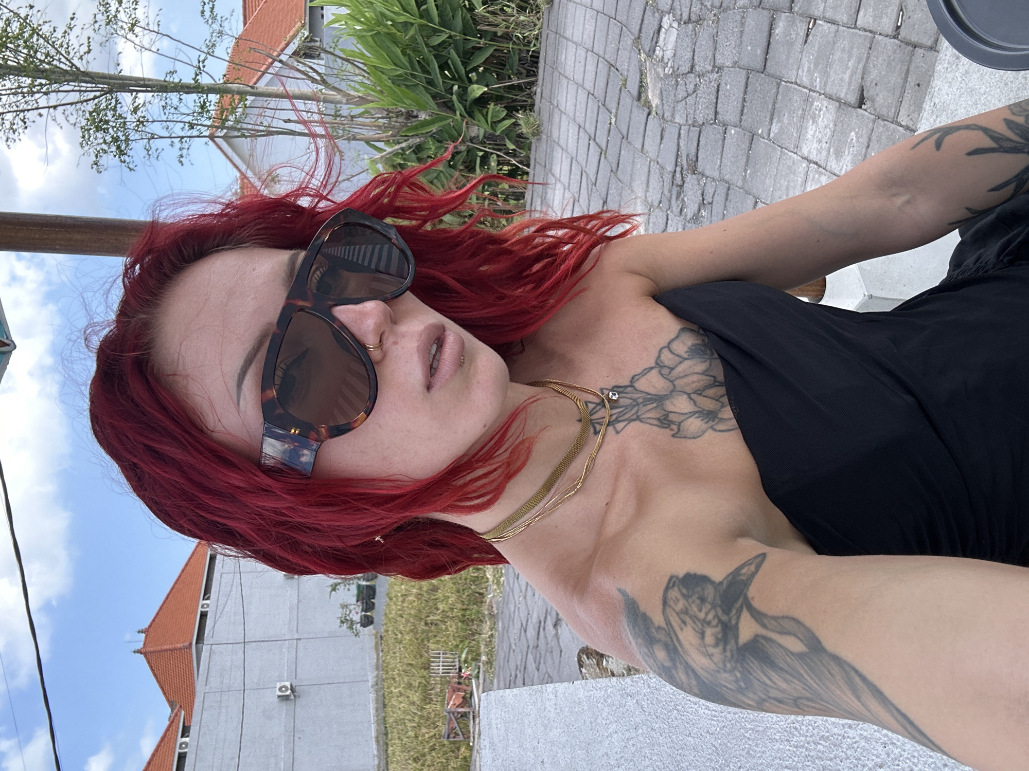 A woman with bright red hair and sunglasses is posing outdoors, likely in North Kuta, Indonesia, given the tropical foliage and distinct architecture. She is wearing a black strapless top and several gold necklaces. Tattoos are visible on her arm and chest, including a large bird tattoo on her left arm and floral designs on her chest. She has a nose ring and a lip piercing. The background features a paved courtyard, lush green plants, and buildings with orange tiled roofs, characteristic of Indonesian architecture. The sky is partly cloudy.