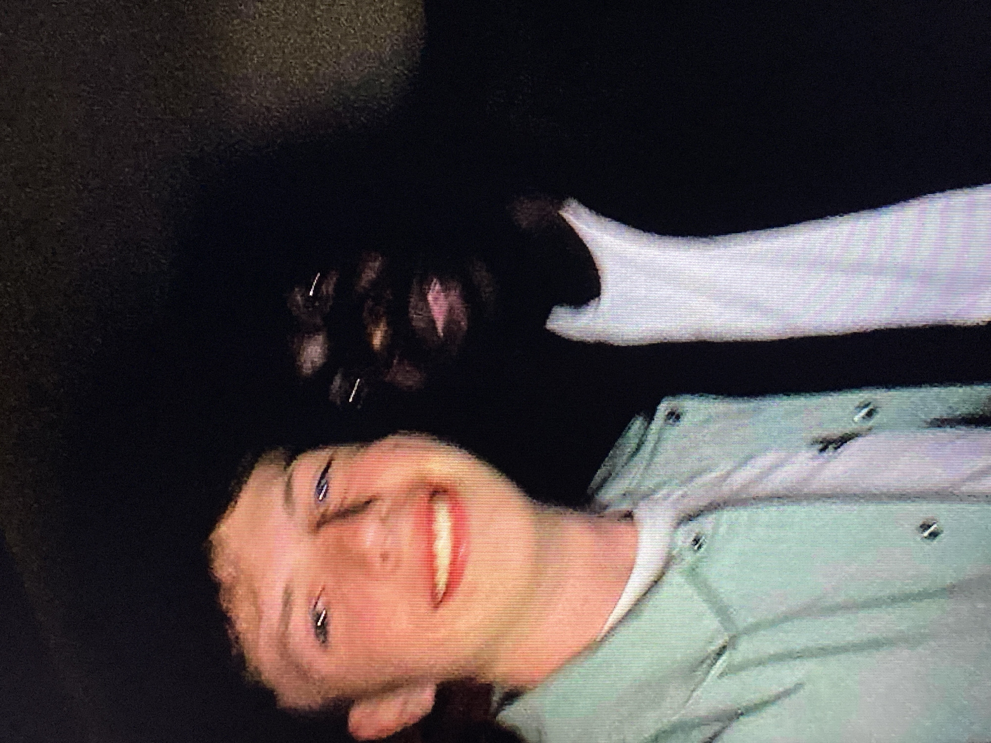 The image is a close-up, selfie-style photograph of two young men. The man on the left is Caucasian with light brown hair, freckles, and a wide smile, wearing a light blue button-up shirt over a white t-shirt. The man on the right is Black with dark skin and is largely obscured by shadow, with only his face and a white t-shirt collar visible. Both men appear to be in a dimly lit environment, possibly indoors or at night, with the background completely dark. The image quality is low, with visible pixelation and noise. There is no discernible urban environment or location information, including anything related to Poznan, Poland.