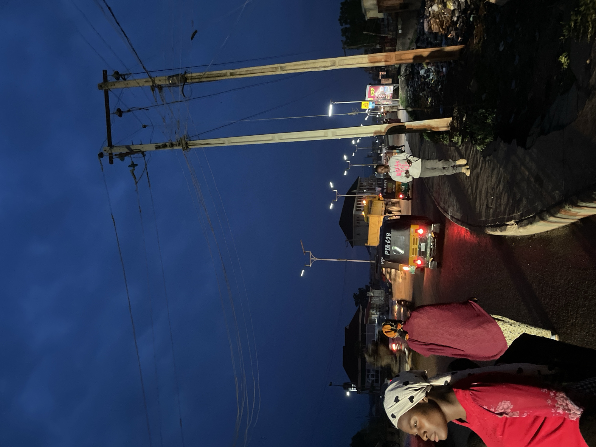 The image captures a nighttime scene on a street in Bukuru, Nigeria. A yellow and black "Keke Napep" (auto-rickshaw) with license plate PTA-698 is parked on the street. Two women, one in a red shirt and white head covering with black hearts, and another wearing a maroon shawl, are standing near the Keke Napep. A man in a white hooded sweater and grey pants stands further down the street. Overhead, power lines and street lights illuminate the area. A pile of garbage is visible along the side of the road. The sky is dark blue.
