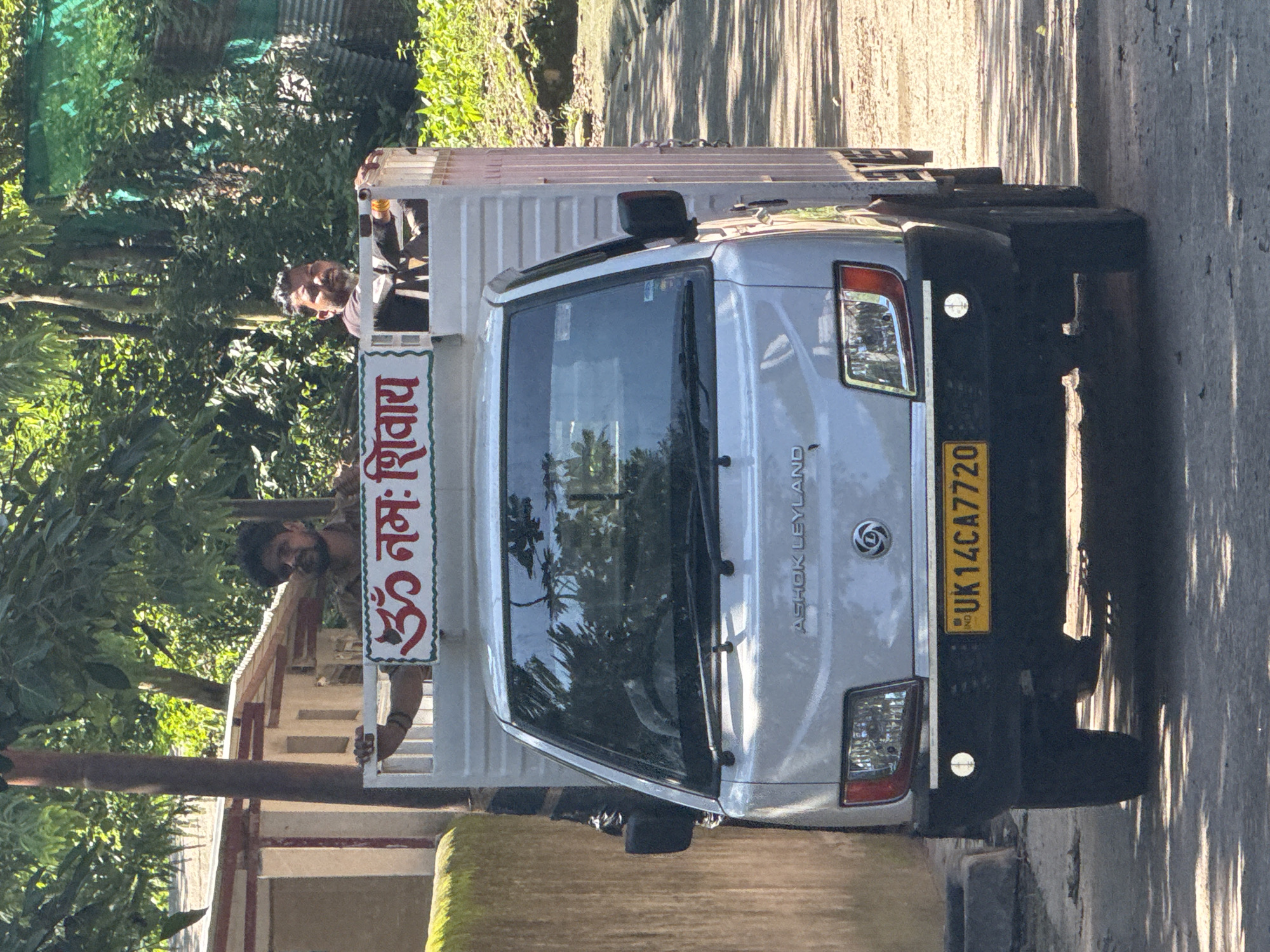 In Rishikesh, India, a white Ashok Leyland light truck is parked on a paved road shaded by trees. Two men are visible in the open back of the truck. The man on the left, wearing a brown shirt, leans on the white railing of the truck. The man on the right, wearing a dark shirt, sits with his hands on the railing. A sign with red Hindi script, "ॐ नमः शिवाय" (Om Namah Shivaya), is affixed to the white railing between the men. The truck's front license plate reads "UK 14CA 7720". The surrounding environment features foliage, a building with a red-trimmed roof, and a wall with green moss. The lighting suggests it is daytime.