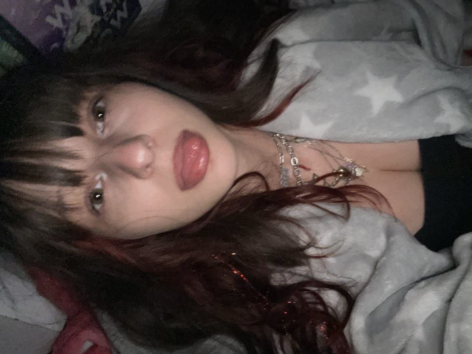 A person with dark hair and bangs is lying down. They have red highlights in their hair and fair skin. The person's lips are parted, and they are wearing necklaces. A gray blanket with white stars is draped over them, revealing a glimpse of a black top. The image is taken at a close-up angle, focusing on the person's face and upper body. The overall lighting is soft and muted.