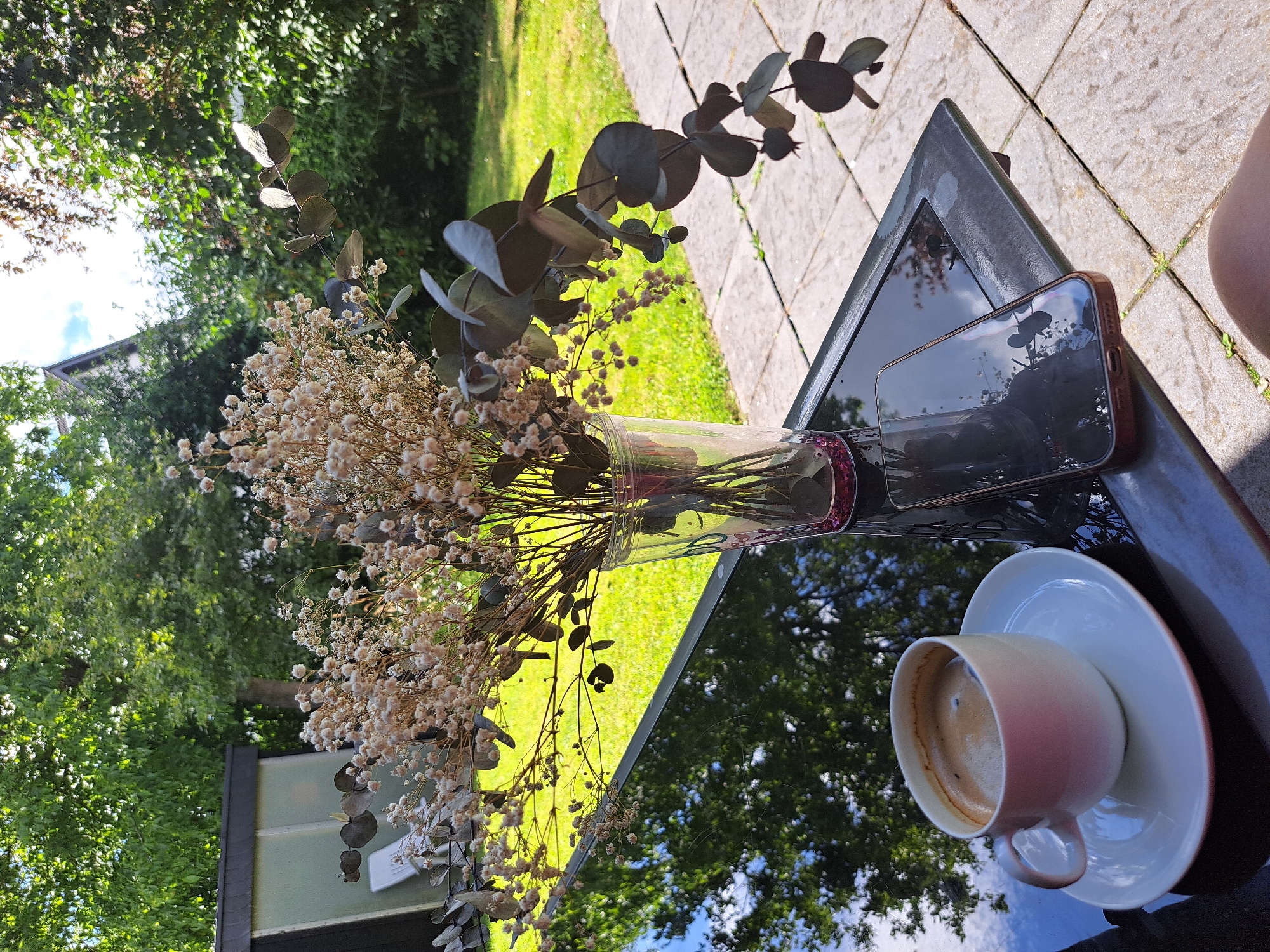 A smartphone, a cup of coffee on a saucer, and a vase of dried flowers are arranged on a black, reflective table. The smartphone is lying face down, showing its metallic back and a camera lens. The cup is a pale pink and white, filled with a dark liquid, likely coffee, with a layer of foam on top. The vase is clear glass and contains a bouquet of delicate white flowers and eucalyptus branches.
The scene is set outdoors on a sunny day. Green grass and trees are visible in the background, some of which are reflected in the glossy surface of the table. The ground is paved with light-colored tiles. The overall impression is one of a peaceful, relaxed moment, perhaps a break in the day. There is no visible text in the image.