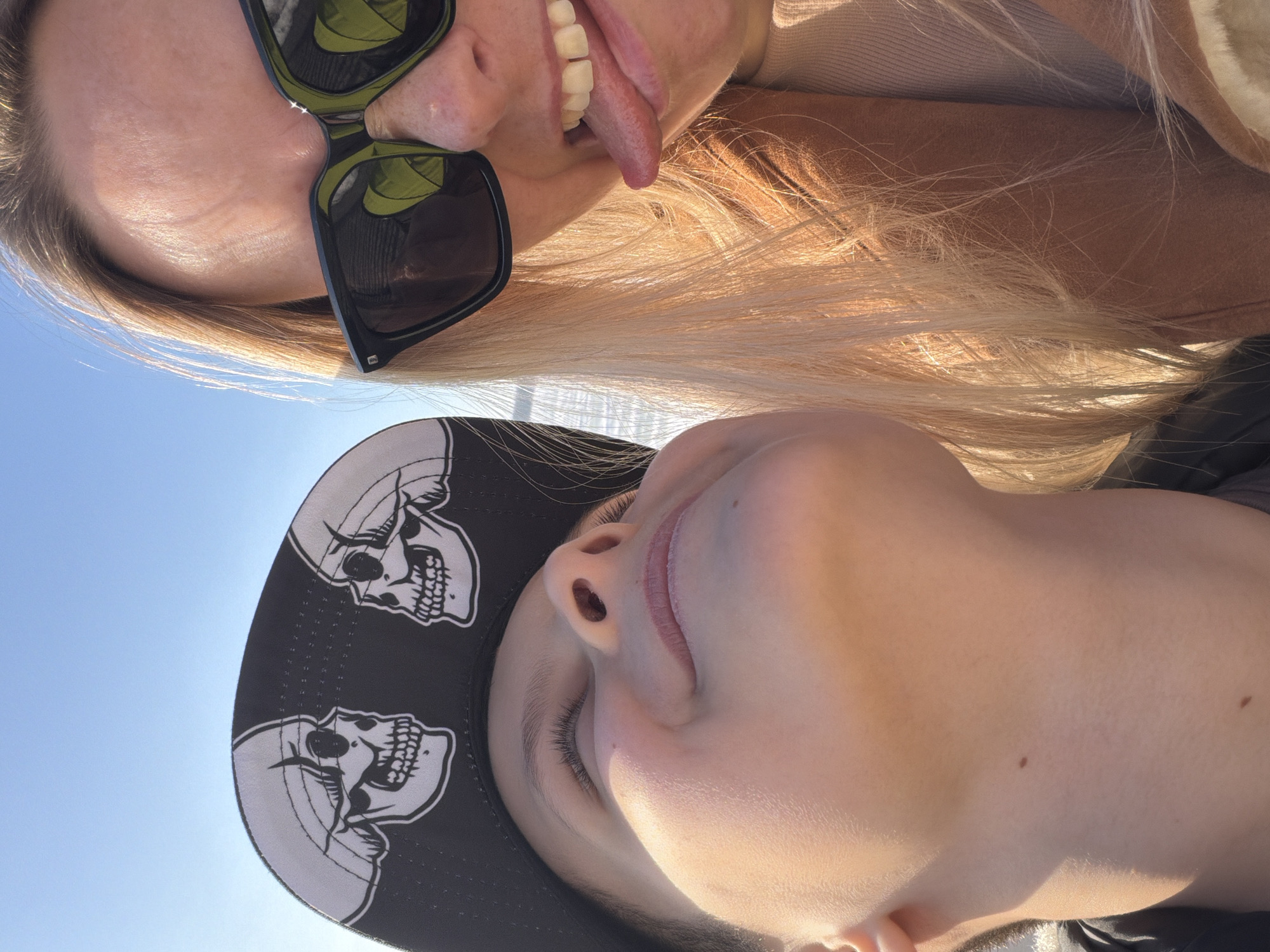 Two individuals are closely positioned, appearing to take a selfie. One person, wearing sunglasses and with blonde hair, is sticking their tongue out. The other person is wearing a baseball cap adorned with skull designs. The background appears to be a clear sky. The specific location in Votkinsk, Russia, is not discernible from the image.
