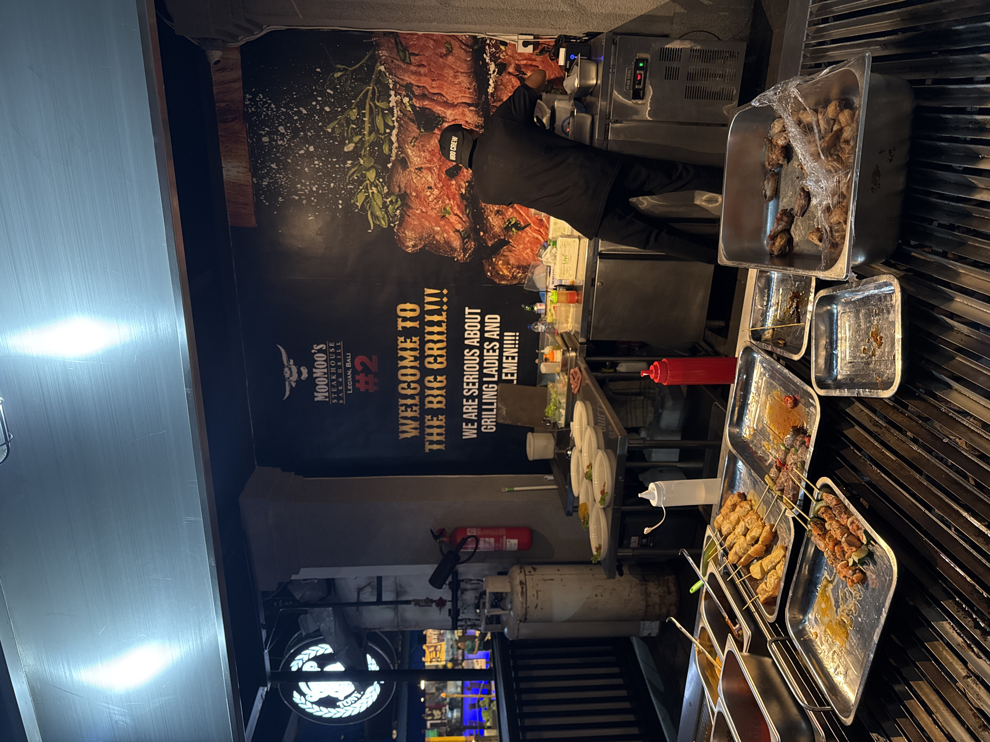 This image depicts an outdoor food stall or restaurant in Kuta, Indonesia, likely a steakhouse and grill based on the signage. The stall features a large backdrop with a graphic of a steak and text that reads "MooMoo's Steakhouse Bar & Grill" and "Welcome to the Big Grill!!! We are serious about grilling ladies and gentlemen!!!!". In the foreground, a grill with multiple trays of prepared skewers and meats is visible, some covered with plastic wrap. A person wearing a black shirt with "MOO CREW" on the cap is actively working at a stainless steel counter with various kitchen equipment, including what appears to be an industrial refrigerator. Other elements present include gas tanks, a fire extinguisher, bottles of sauces or condiments, and plates set up for service. The overall scene suggests a busy nighttime operation at a food establishment.