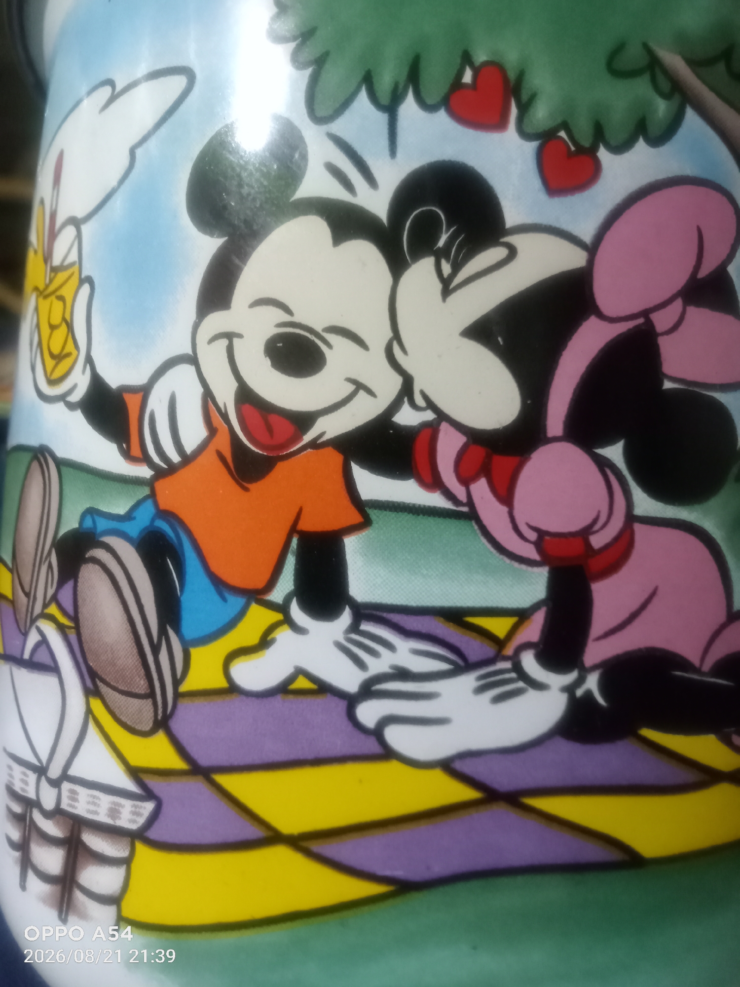 The image is a close-up of a mug featuring a cartoon depiction of Mickey Mouse and Minnie Mouse. They are shown in a romantic pose under a tree with red hearts hanging from its branches. Mickey, wearing an orange shirt and blue shorts, is holding a drink and smiling, with his arm around Minnie. Minnie, dressed in a pink dress and bow, is leaning her head on Mickey's shoulder. They are sitting on a picnic blanket with a checkered pattern of yellow and purple. The background is a light blue sky with green grass at the bottom. The bottom left corner of the image displays a timestamp "OPPO A54 2026/08/21 21:39", indicating the photograph was taken with an OPPO A54 phone on August 21, 2026, at 21:39. There is no information in the image that suggests a location in Jambi, Indonesia.