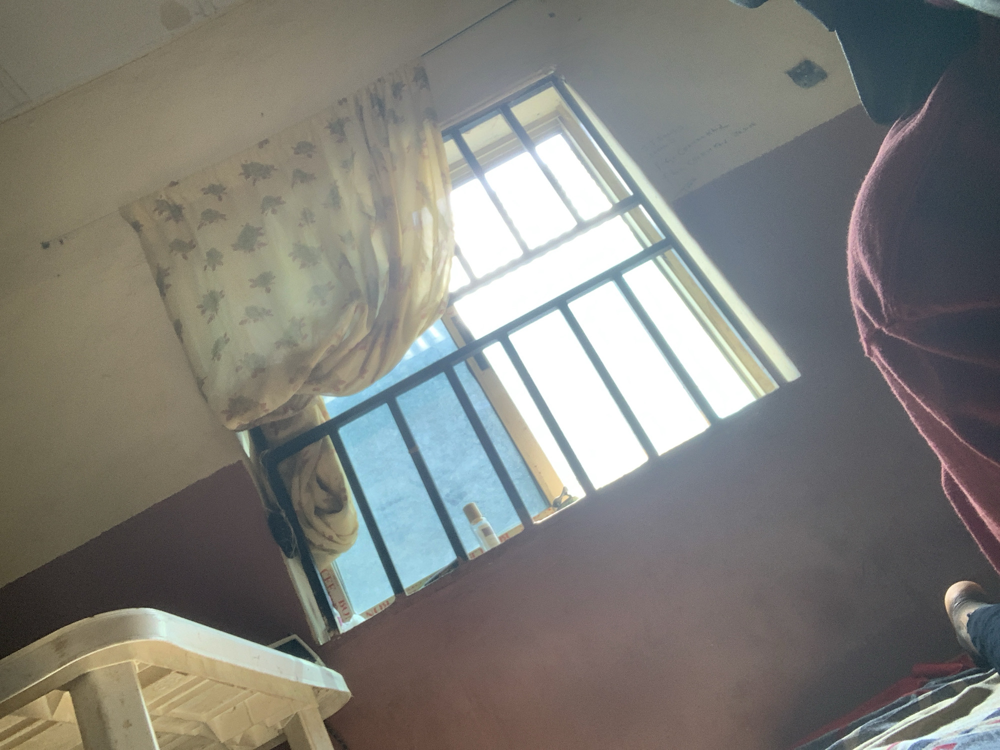 The image depicts a view from inside a room, looking upwards towards a window. The window has a metal security grill and is letting in bright sunlight. A light-colored curtain with a floral pattern is partially visible, drawn across part of the window. The walls of the room appear to be painted in muted colors. In the foreground, a person's feet, clad in dark socks, are visible, and a portion of a dark red blanket or fabric is also in view, suggesting someone is lying down or sitting. The scene suggests a domestic environment. While the location is stated as Jauro Yinu, Nigeria, there are no specific visual cues within the image that definitively confirm this geographical location.