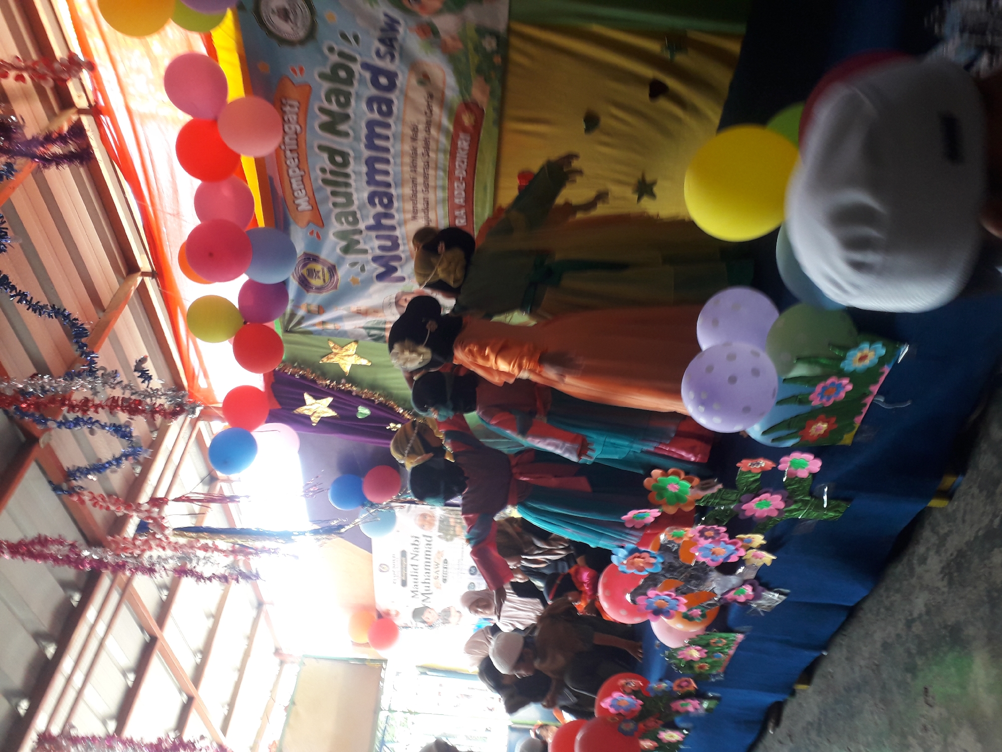 The image captures an indoor event, likely a celebration or ceremony, taking place at an unspecified location in Cankuang, Indonesia. The scene is decorated with colorful streamers and balloons hanging from the ceiling and arranged around the venue. A banner in the background reads "Memperingati Maulid Nabi Muhammad SAW" (Commemorating the Birthday of Prophet Muhammad SAW), indicating the nature of the event.
In the foreground, a group of children, dressed in traditional attire, are gathered on a stage or raised platform. Some of the children are wearing brightly colored dresses and headscarves, while others are wearing white caps. They appear to be participating in a performance or presentation, with some children facing away from the camera.
The environment appears to be somewhat rustic, with exposed ceiling beams and what looks like a makeshift stage covered in a blue cloth. The lighting is bright, suggesting an indoor setting with ample natural or artificial illumination. The overall atmosphere seems festive and communal, with a focus on religious observance and cultural celebration.