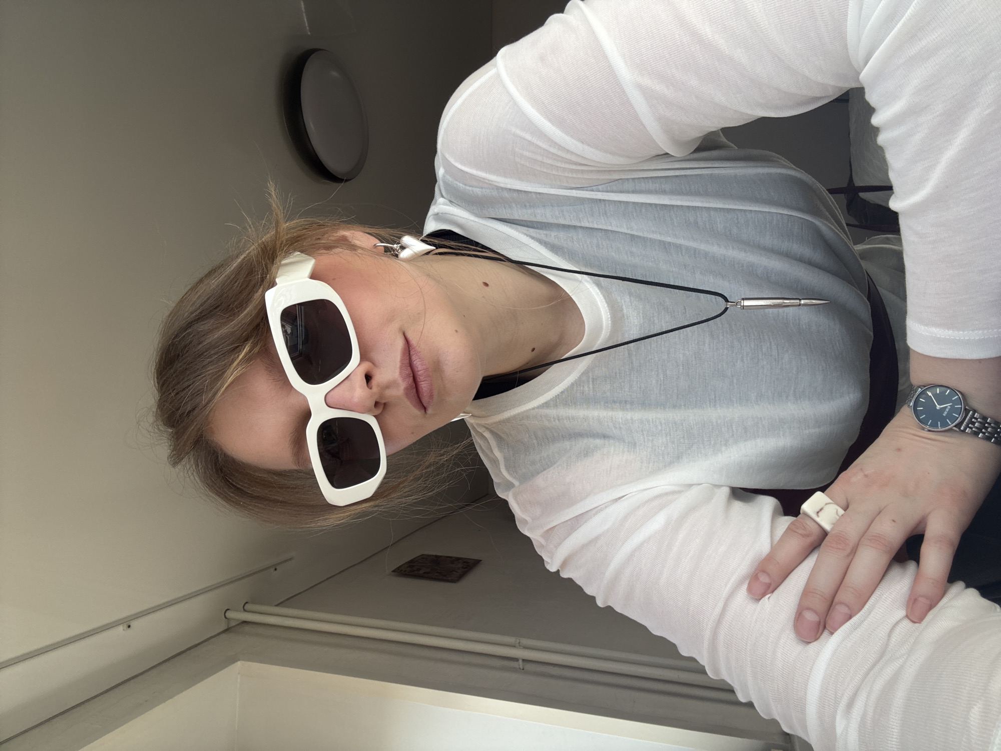 A fair-skinned person with light brown hair is looking down at the camera, wearing large white-framed sunglasses. The person wears a long-sleeved white shirt over what appears to be a black underlayer. A silver, pen-shaped pendant hangs from a black cord around their neck. A silver wristwatch is on their left wrist and a large, square, off-white ring adorns their left ring finger. The background includes a gray wall with a circular light fixture and a glimpse of a white door frame or corner. The image orientation is rotated to the left. There is no clear evidence indicating the location to be Izhevsk, Russia.