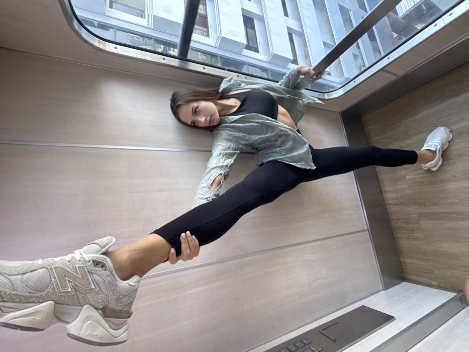 A young adult female stands inside a panoramic glass elevator within the Mahanakhon SkyWalk at King Power Mahanakhon Tower in Bangkok, Thailand. She has long, dark brown hair and is wearing a black ribbed sports bra, an unbuttoned light green/grey long-sleeved shirt, and black ribbed leggings. Her right leg is extended vertically upwards, with her right hand gripping her calf/ankle. A beige New Balance 9060 sneaker is visible on her raised right foot. Her left foot, wearing a white chunky sneaker, rests on the light wood-grain floor. Her left hand grips a silver handrail positioned along the curved observation window. Through the window, a cityscape of modern high-rise buildings and urban structures is visible. The elevator interior features light wood-paneled walls with silver vertical seams and base trim, and a silver control panel is present on the left wall.