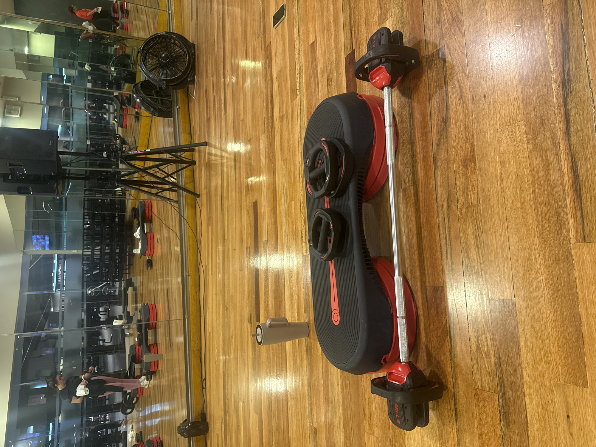 The image shows a fitness studio in Puebla, Mexico, with mirrored walls reflecting exercise equipment and people exercising. A Les Mills SMARTBAR™ weight training system lies on the wooden floor. The SMARTBAR™ comprises a barbell, weight plates with handles, and clips to secure the plates.