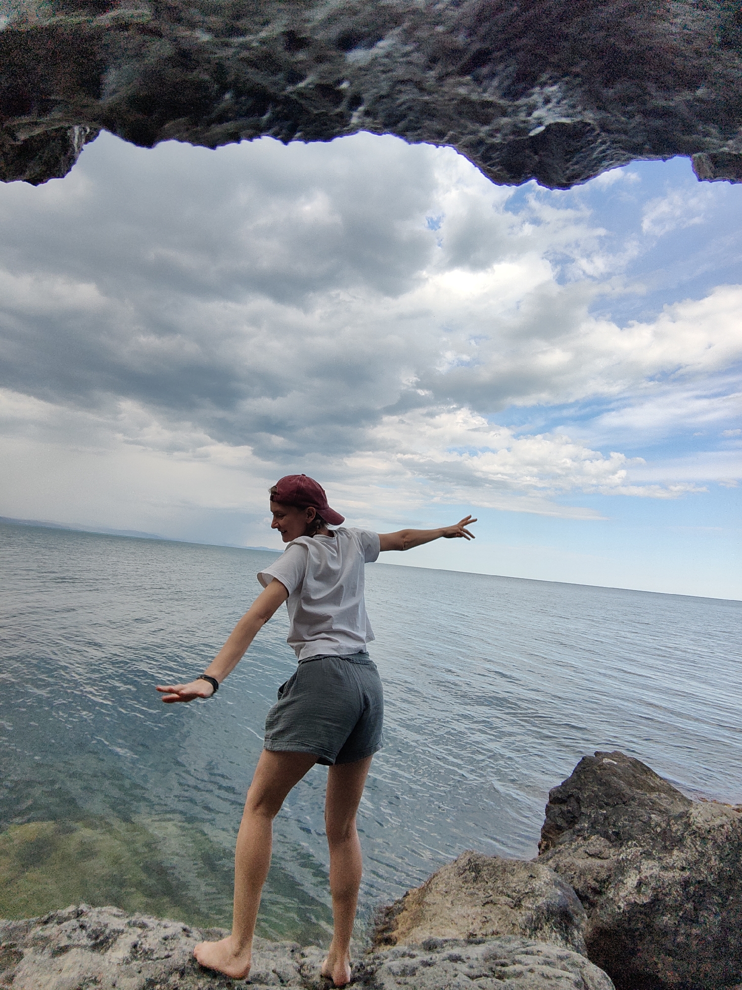 The image captures a person standing on rocky terrain near the sea. The person is facing away from the camera and towards the water, with their arms outstretched as if balancing. They are wearing a white t-shirt, grey shorts, and a red baseball cap. The sea extends to the horizon, meeting a cloudy sky. The foreground is dominated by rough, grey rocks, some of which appear to be partially submerged in the clear water. The overall scene suggests an outdoor, natural environment. The location is stated to be in the city of Utios, Ukraine.
