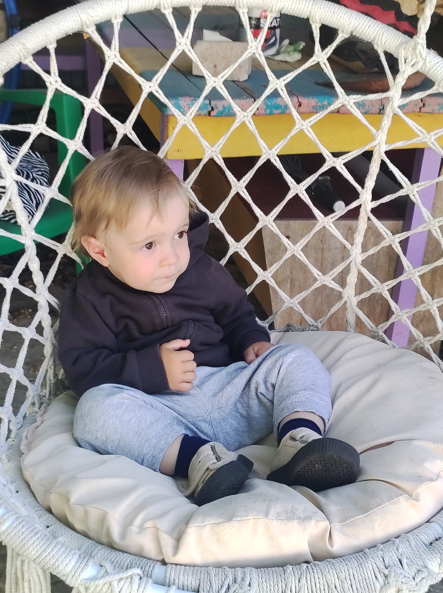 A young toddler with light brown hair and brown eyes is seated in a white, macrame-style hanging chair. The child wears a dark, possibly black or dark grey, hooded zippered sweatshirt, light grey sweatpants, dark blue socks, and light-colored shoes with thick, dark soles. The child is looking towards the right side of the frame with a calm, curious, or thoughtful expression, with their right hand resting on their knee.
The hanging chair features a cream-colored cushion where the child is sitting. Behind the chair, the background reveals a rustic, multi-colored structure, possibly a table or shelf, with visible sections painted in green, yellow, purple, and blue. Some of the painted surfaces appear worn or cracked, suggesting an outdoor or well-used indoor setting. A black and white striped object is partially visible on the left side of the background. On the upper right side of the background, a small portion of a black object with a white label is visible, displaying the partial text "STAT".
The lighting suggests it is daytime. The overall scene conveys a relaxed and informal atmosphere.