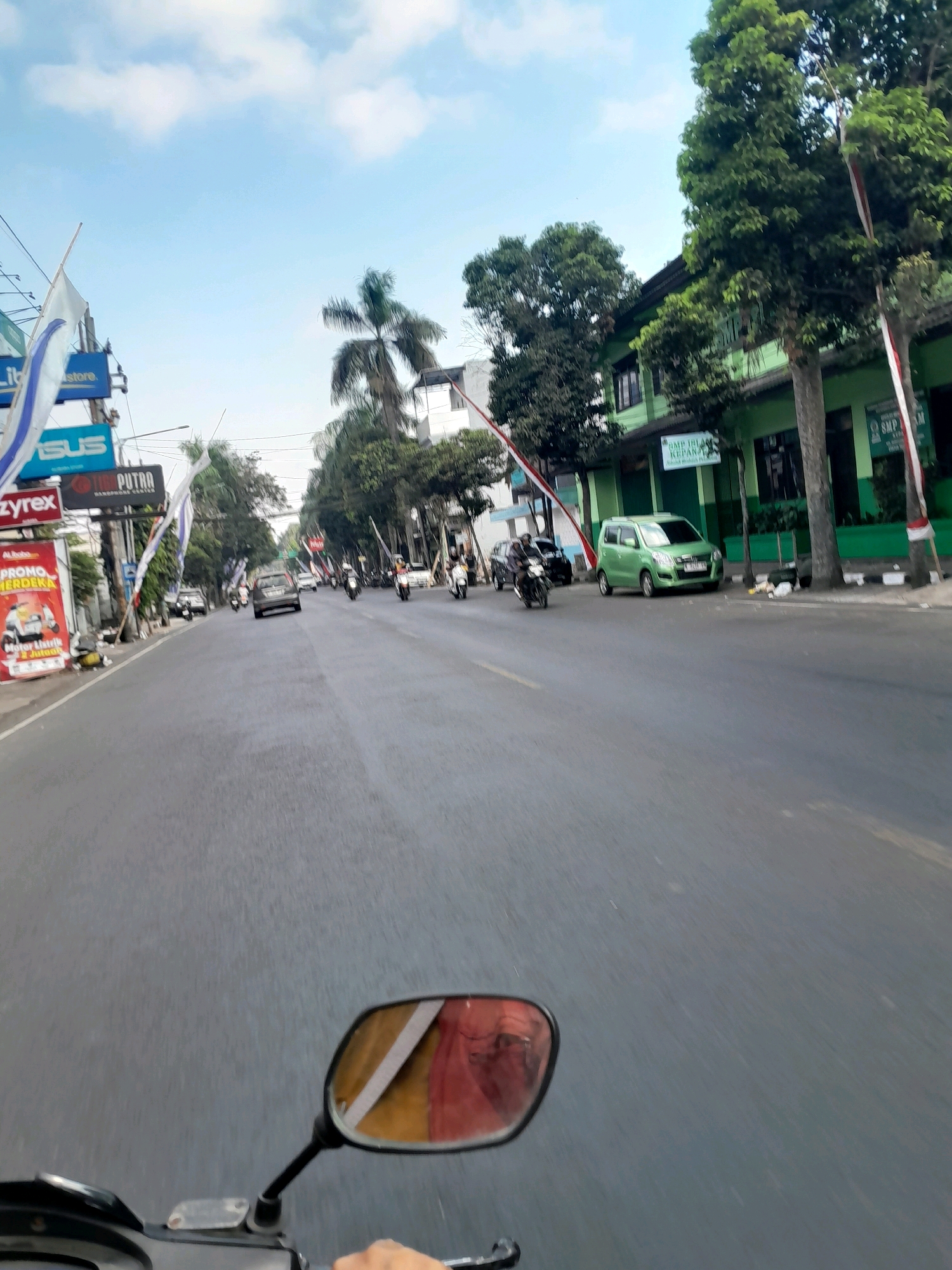 The image captures a street scene in Kepanjen, Indonesia. The perspective is from a moving vehicle, likely a motorcycle, as indicated by the visible handlebars and a rearview mirror reflecting a person wearing red clothing. The road is asphalt, with lane markings visible. On the left side of the road, there are buildings with signs, including one for "ASUS". There's also a red advertisement banner for "PROMO MERDEKA" featuring a scooter. Further down the road, various vehicles, including cars and motorcycles, are in motion. Palm trees and other trees line the street, and a building with green siding and windows is visible on the right. Red and white flags are displayed along the street, possibly indicating a celebration or event. The sky is partly cloudy, with a bright blue expanse. The overall impression is that of a typical day in a bustling town in Indonesia.
