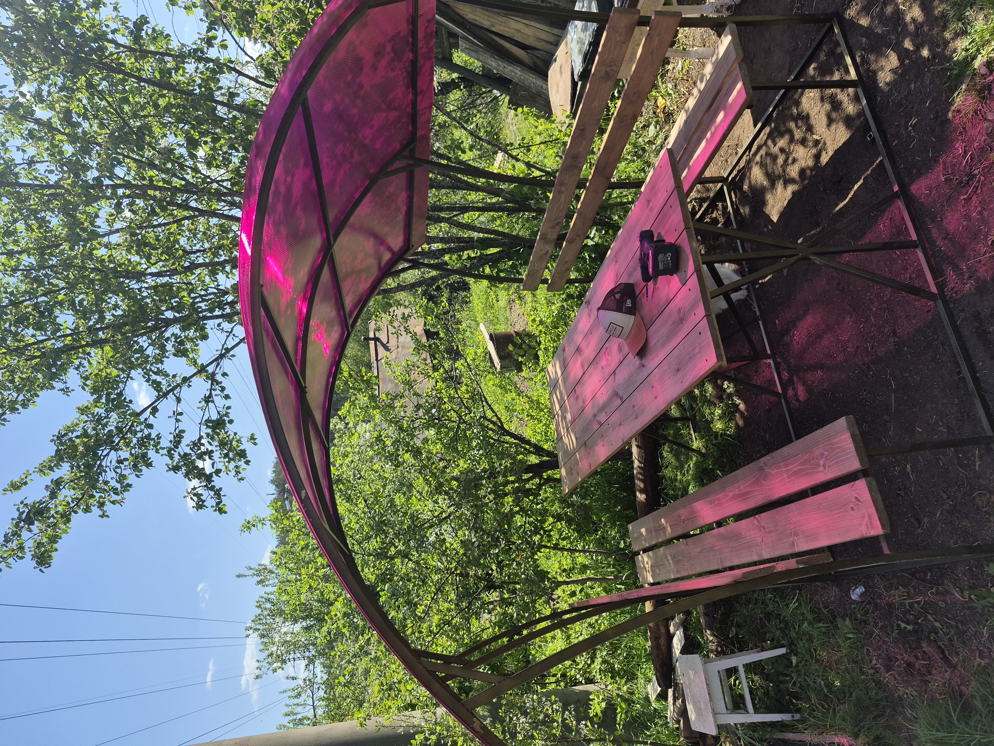 The image depicts an outdoor scene with a picnic table and benches under a curved, translucent, magenta-colored canopy. The table is made of wooden planks, and there is a hat and a black object (possibly a tool) on it. The canopy is supported by a metal frame. The area around the table appears to be a yard or garden, with trees, foliage, and bare ground. Power lines are visible against a blue sky in the background. A white chair is located next to the table. The location is uncertain.