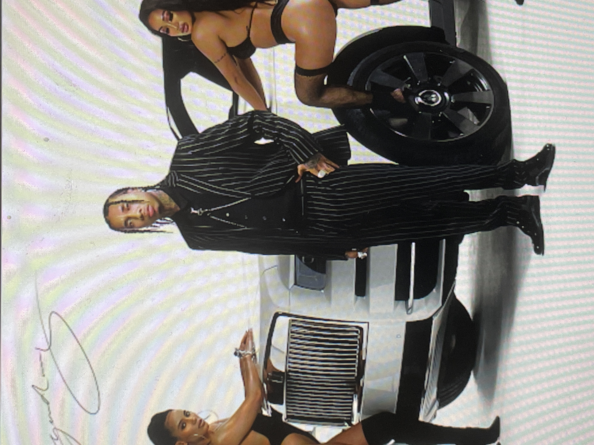 The image displays a promotional photoshoot featuring one man and two women posing with a white luxury SUV, likely a Rolls-Royce Cullinan, against a light-colored studio background. The overall composition is dynamic, with the subjects arranged around the vehicle.
The central figure is a man with light brown skin and braided hair, wearing a black pinstriped suit over a dark vest and white shirt, accessorized with a chain necklace and rings. He stands confidently, looking towards the camera, with one hand near his waist and the other slightly raised. He wears black boots.
Two women are positioned on the SUV. One woman with long dark hair and light brown skin is draped over the rear wheel and fender, wearing a black bikini-style top, matching bottoms, and thigh-high black stockings. She gazes towards the camera. The second woman, with similar features, lies on the front hood and grille of the SUV, also dressed in a black bikini-style top and bottoms, with one arm raised in a pose.
The white SUV serves as a prominent prop, featuring black multi-spoke wheels and a distinctive chrome grille. The background is a plain, light studio wall, exhibiting subtle vertical banding, possibly due to lighting or screen capture.
The activity taking place is a stylized photoshoot, possibly for music album art or an advertisement, conveying an opulent and edgy aesthetic. The subjects project confidence and allure. In the bottom left corner of the image, a faint, stylized signature, possibly "Tyga," is visible. While the image itself doesn't offer specific location cues beyond a studio setting, the provided context indicates the shoot took place in Sofia, Bulgaria.