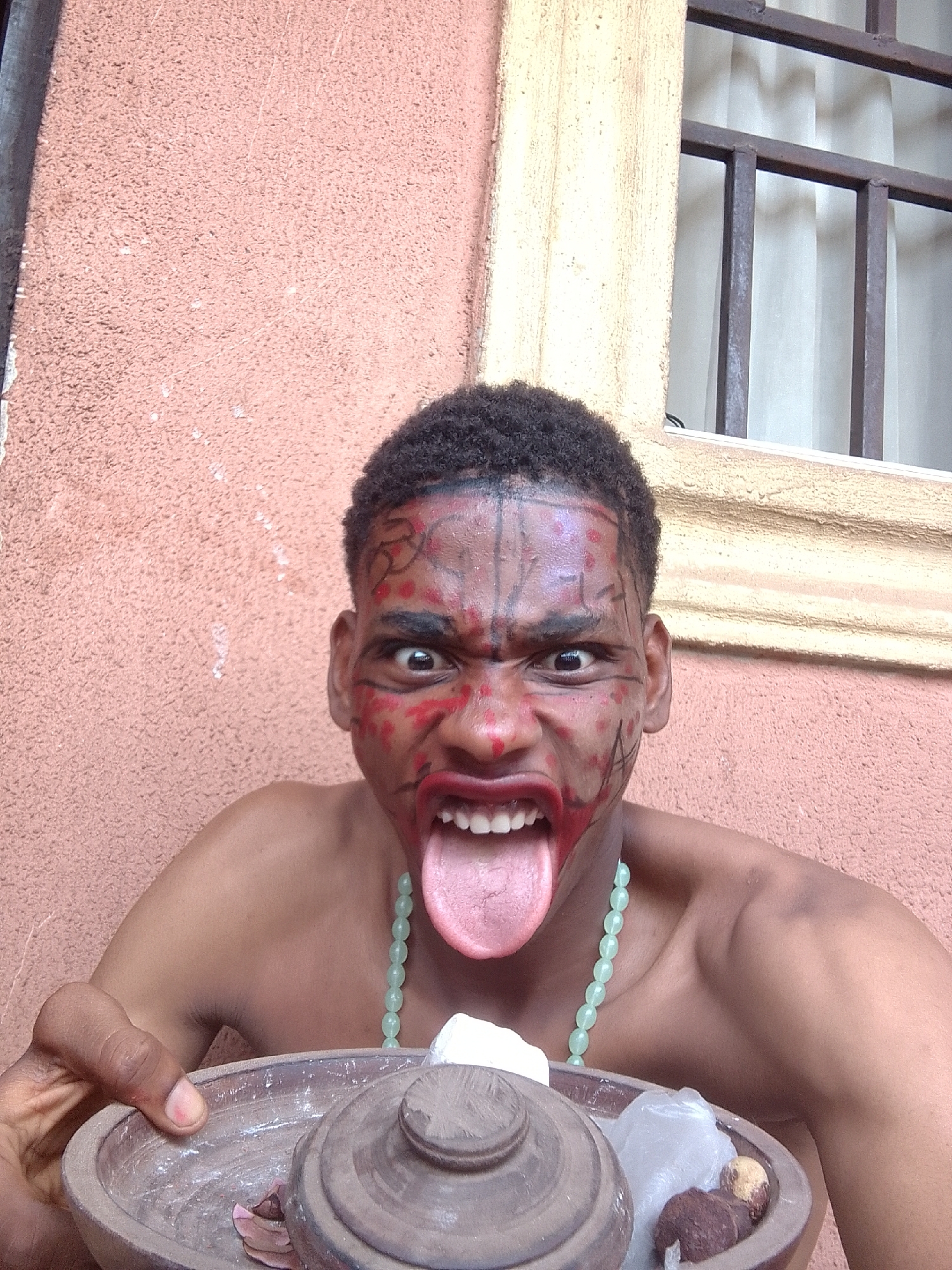 A person, likely a young man with dark skin and short, dark curly hair, is captured in a close-up, chest-up shot. Their face is painted with striking red and black patterns, including lines across the forehead, around the eyes, on the cheeks, and covering the lips. The individual wears a light green beaded necklace. They are making an exaggerated, intense facial expression with wide, bulging eyes, a furrowed brow, and an open mouth with the tongue sticking out, conveying a fierce or playful demeanor.
The person holds a shallow, round, dark brown wooden or ceramic bowl in their left hand. The bowl contains various items, including a matching lid partially covering white, irregular-shaped lumps (possibly chalk or a similar substance), a small lumpy brown object, and some red or brown flakes.
The background features a textured, reddish-brown wall. To the upper right, a window with dark metal security bars and light-colored curtains behind them is visible, suggesting an indoor or semi-outdoor setting like a porch or courtyard. The lighting appears bright and even, indicating daytime. There is no visible text in the image.