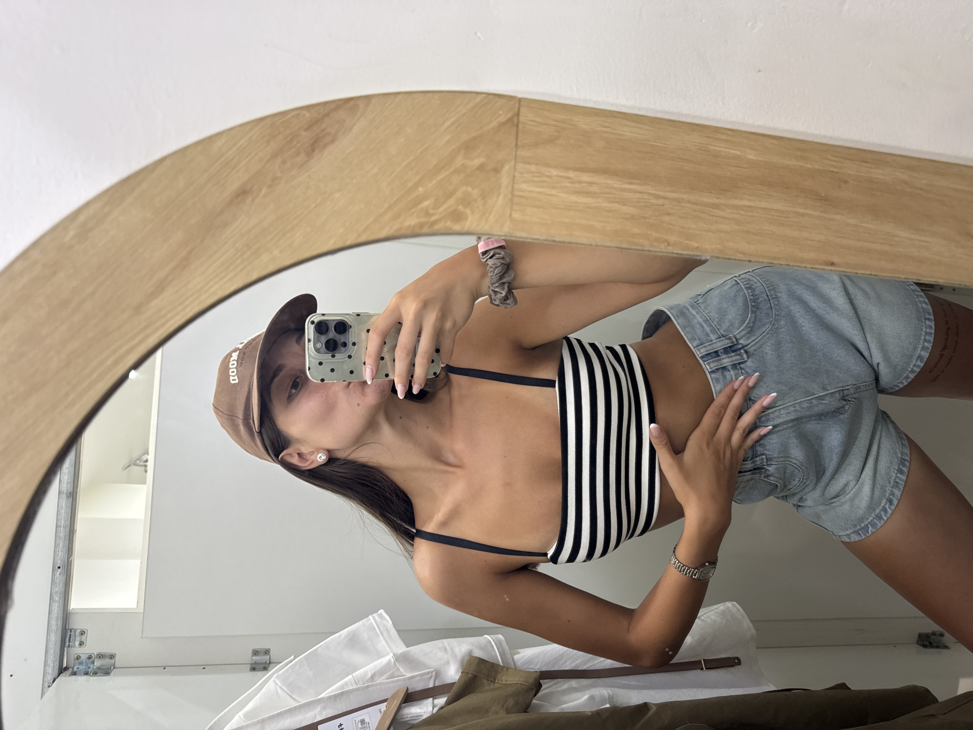A woman wearing a brown baseball cap, a black and white striped crop top, and light blue denim shorts is taking a mirror selfie. She is holding a phone with a spotted case. The mirror has a light wood frame. In the background, there is a white wall and some clothing items. The location cannot be determined from the image.