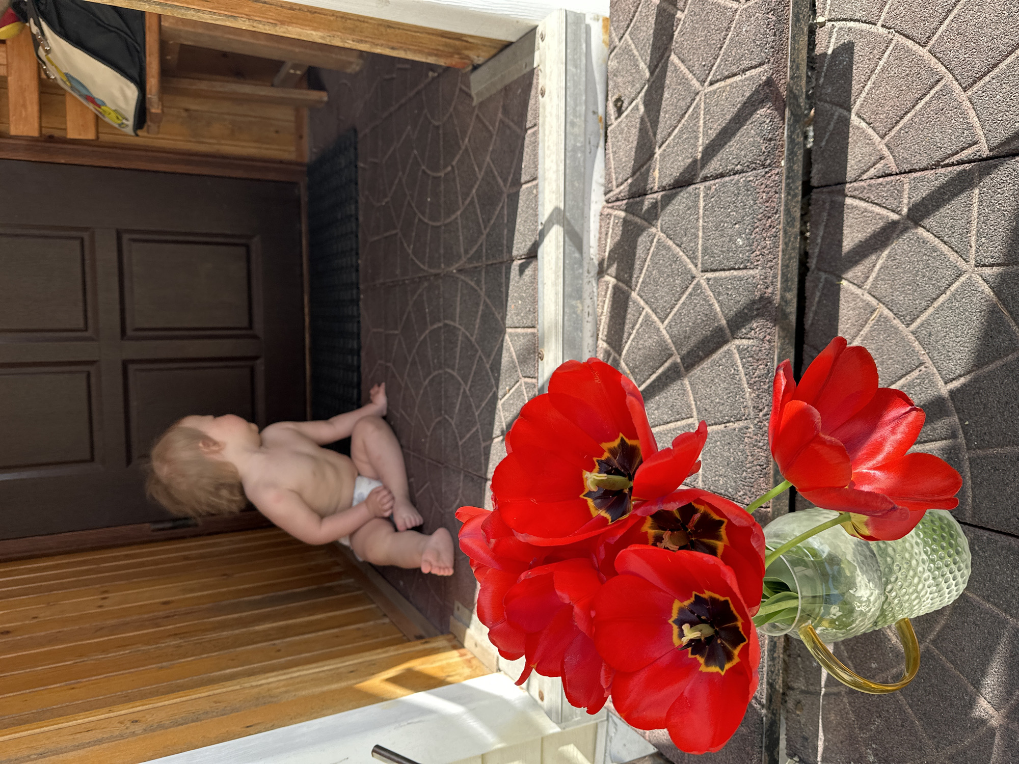 A baby lies on a wooden floor just inside an open doorway. The baby, wearing only a diaper, is fair-skinned with light hair. A brown door and wooden slats extend into the background. Adjacent to the doorway is a wall constructed of gray brickwork in a circular pattern. In the foreground, a bouquet of red tulips is displayed in a green glass pitcher with a yellow handle. The entire scene is bathed in sunlight, creating distinct shadows. The location is in Nizhny Novgorod, Russia.