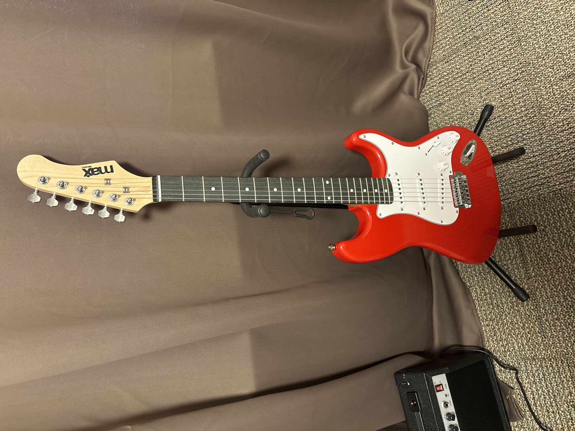 A red electric guitar stands upright on a black guitar stand in front of a brown curtain. The guitar has a light-colored neck with six tuning pegs on the headstock, and a white pickguard on the body. Below the guitar, a small black amplifier with silver knobs and a red power switch is visible. The guitar and amplifier are placed on a textured carpeted floor. The scene appears to be indoors, likely a practice space or home. There is no discernible location in Heerlen, Netherlands mentioned or depicted.