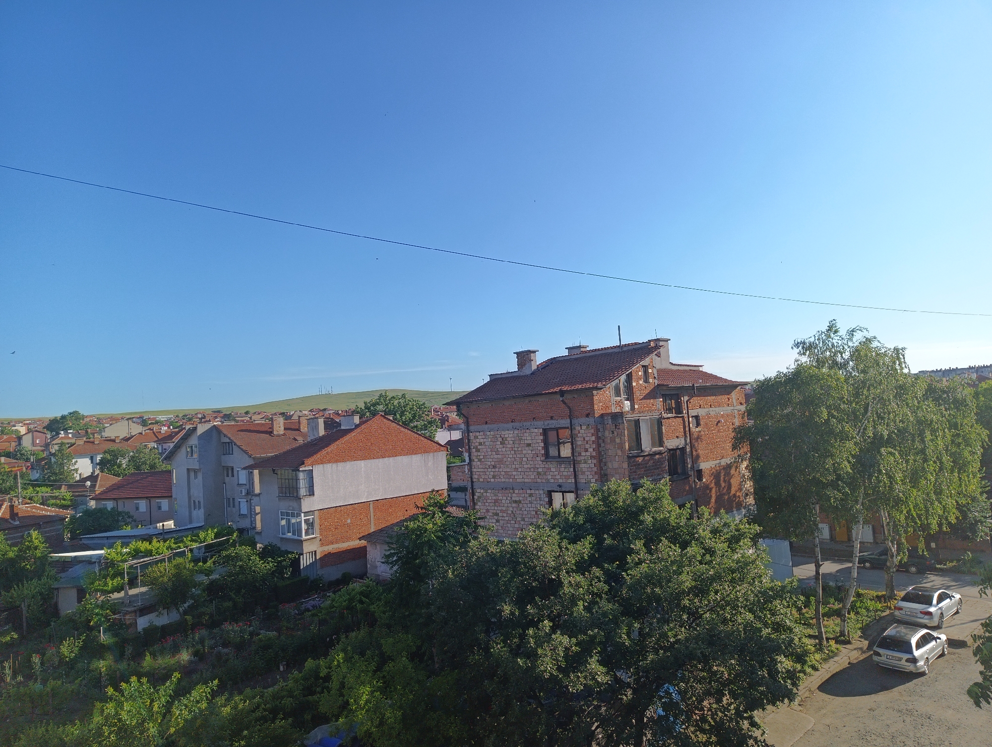 The image shows a residential area in the city of Karnobat, Bulgaria, under a clear blue sky. The foreground features lush green trees and vegetation, suggesting a well-maintained garden or park area. Behind this greenery, several houses are visible, characterized by red-tiled roofs and brick exteriors. One prominent building in the mid-ground appears to be under construction or renovation, with exposed brickwork and unfinished sections. Further in the background, a sprawling landscape of similar houses stretches towards rolling hills, indicating a typical suburban or semi-rural setting within the city. Two silver cars are parked on a paved road in the right side of the image, adjacent to the houses and trees. No people are visible in the scene, and no specific actions or interactions are depicted. The overall impression is a peaceful, sunlit day in a Bulgarian neighborhood.