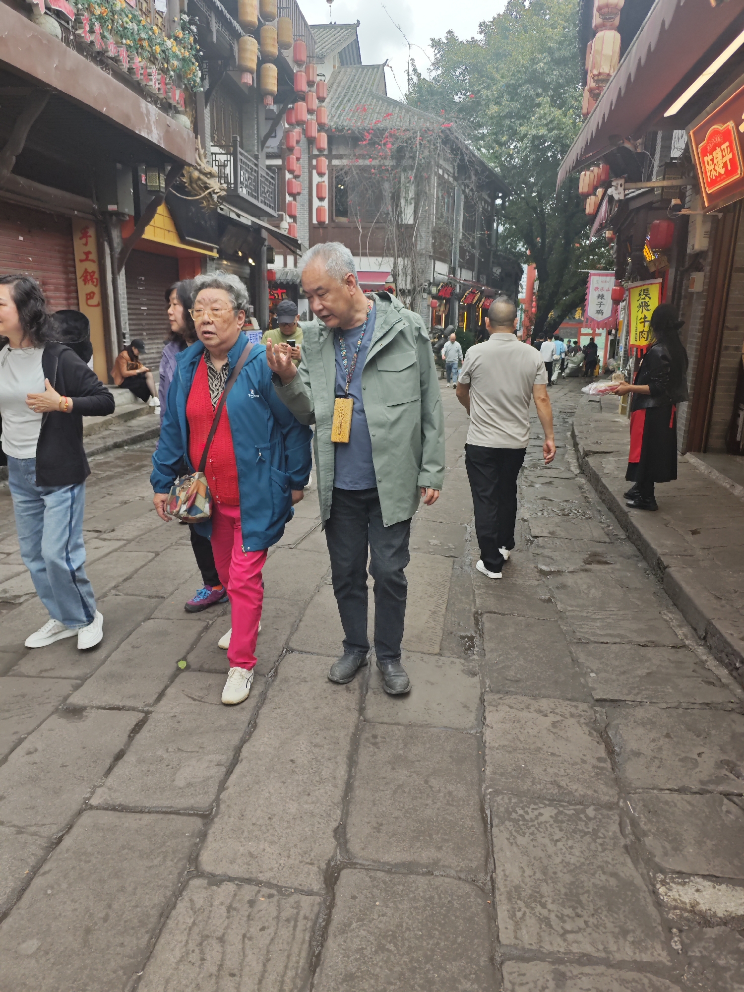 The image captures a street scene in Shapingba District, China. The narrow pedestrian street is paved with large stone blocks. A group of people is walking along the street, including an elderly couple who are the most prominent figures in the foreground. The woman is wearing a bright blue jacket over a red sweater, paired with pink pants and white sneakers. The man is wearing a light green jacket, dark pants, and gray shoes, and has a lanyard around his neck. He appears to be gesturing with his hand as if explaining something. Other pedestrians are present, including a person walking away from the camera with a shaved head and a woman standing in front of a storefront holding a tray. The buildings lining the street have traditional Chinese architectural elements, and there are hanging lanterns and signs in Chinese characters. The weather appears to be overcast, with a gray sky visible above the buildings.