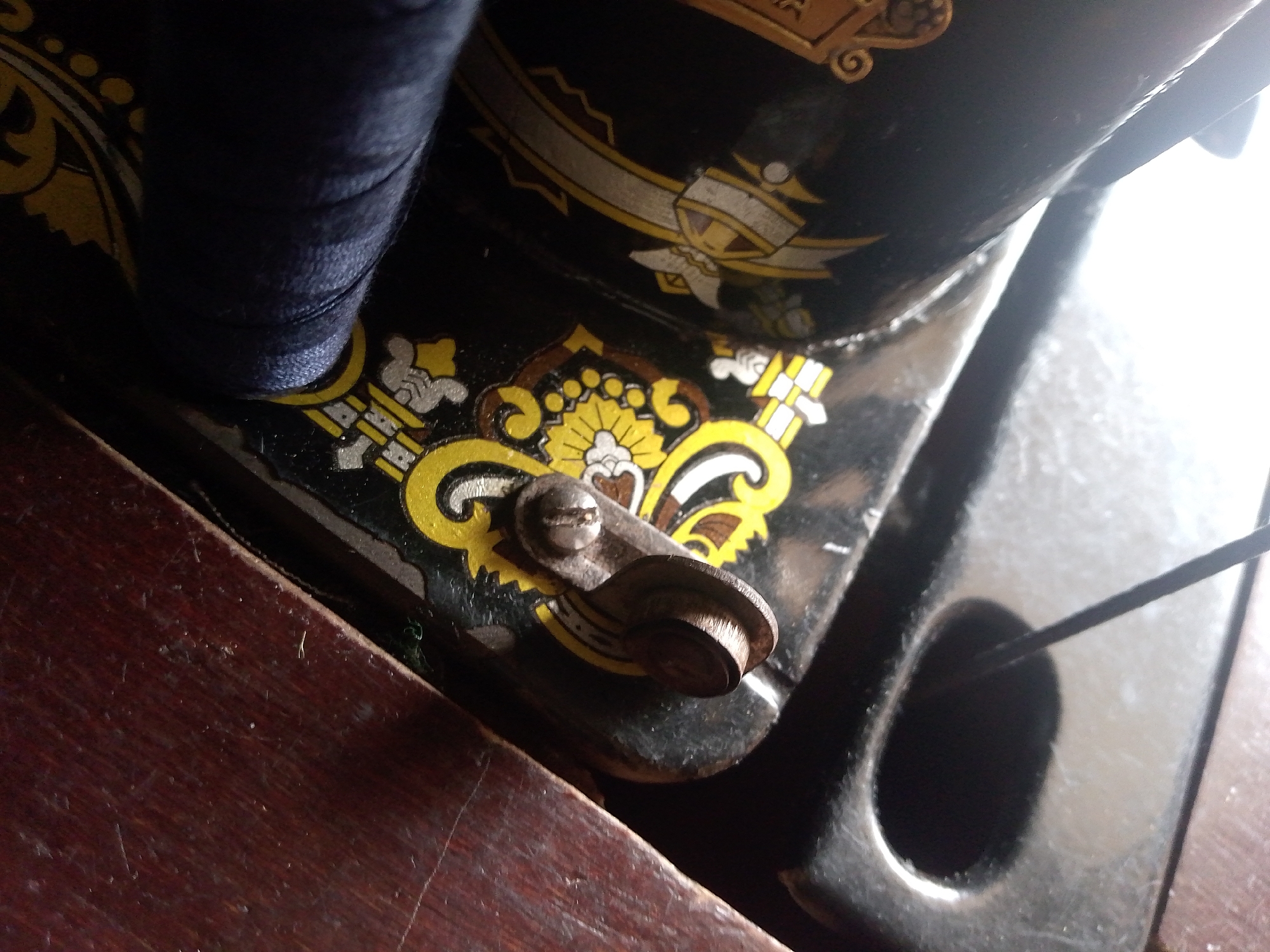 An extreme close-up shot, captured in Jauro Yinu, Nigeria, focuses on a dark blue spool of thread situated on a vintage sewing machine. The sewing machine exhibits ornate yellow and white floral decals against its black metallic surface. A small, silver metal lever with a screw head is prominently visible on the machine, near the thread spool. The machine itself rests on a dark, wooden surface, possibly a table, which shows signs of wear and tear. A thin black string or thread extends from the right side of the frame towards the center, disappearing into a dark opening on the sewing machine's base. The lighting in the scene is uneven, with a bright area towards the upper right, suggesting the presence of a light source or window. No people are visible in the frame.