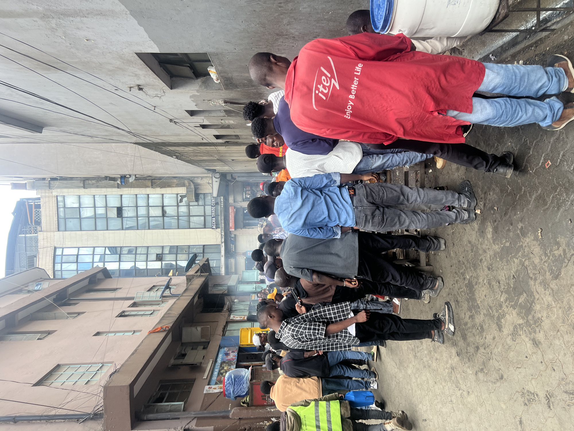 The image depicts a crowded urban street scene, likely in a commercial area of Nairobi, Kenya. The perspective is from behind a line of people, predominantly men, standing and facing away from the viewer.
In the immediate foreground, a man wearing a bright red jacket with the brand "itel" and the slogan "Enjoy Better Life" printed on the back stands next to a large white barrel with a blue lid, seemingly interacting with its contents. He is dressed in light blue jeans and brown shoes. A queue of other men extends behind him, wearing various casual attire including t-shirts, plaid shirts, jeans, and sneakers. One individual towards the left wears a high-visibility yellow vest with the numbers "07407328" visible.
The background reveals a dense urban environment with multiple-story buildings. One prominent building has "ACCRA TRADE CENT" (partially visible) written on its facade, and another sign below reads "DIMAL LIQUOR." The buildings feature numerous windows, some reflecting the sky. Overhead, a network of utility wires crisscrosses the frame. The ground is a worn, dark concrete or asphalt surface. The lighting suggests it is daytime, possibly overcast, creating diffuse shadows. The scene captures a moment of everyday life in a bustling city, with people gathered in what appears to be a line or waiting area.