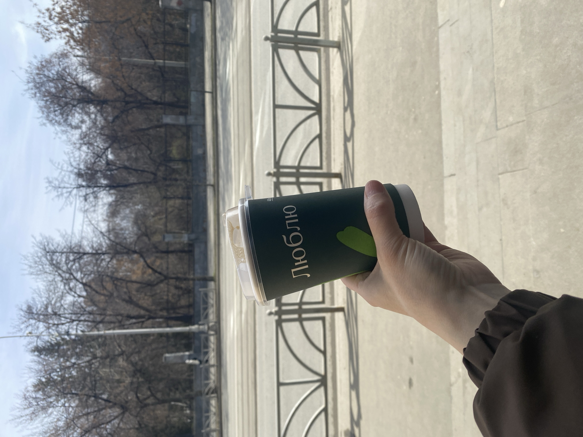 A person holds a green paper cup with the Russian word "Люблю" (I love) written on it. The cup contains a beverage with a light-colored foam visible through the clear plastic lid. The person is wearing a brown jacket or coat.
The background shows a city street with a gray metal fence and a concrete walkway. Bare trees are visible in the distance, suggesting it is late fall or early spring. The sky is pale blue. The scene appears to be in Yekaterinburg, Russia, based on the Russian text on the cup.