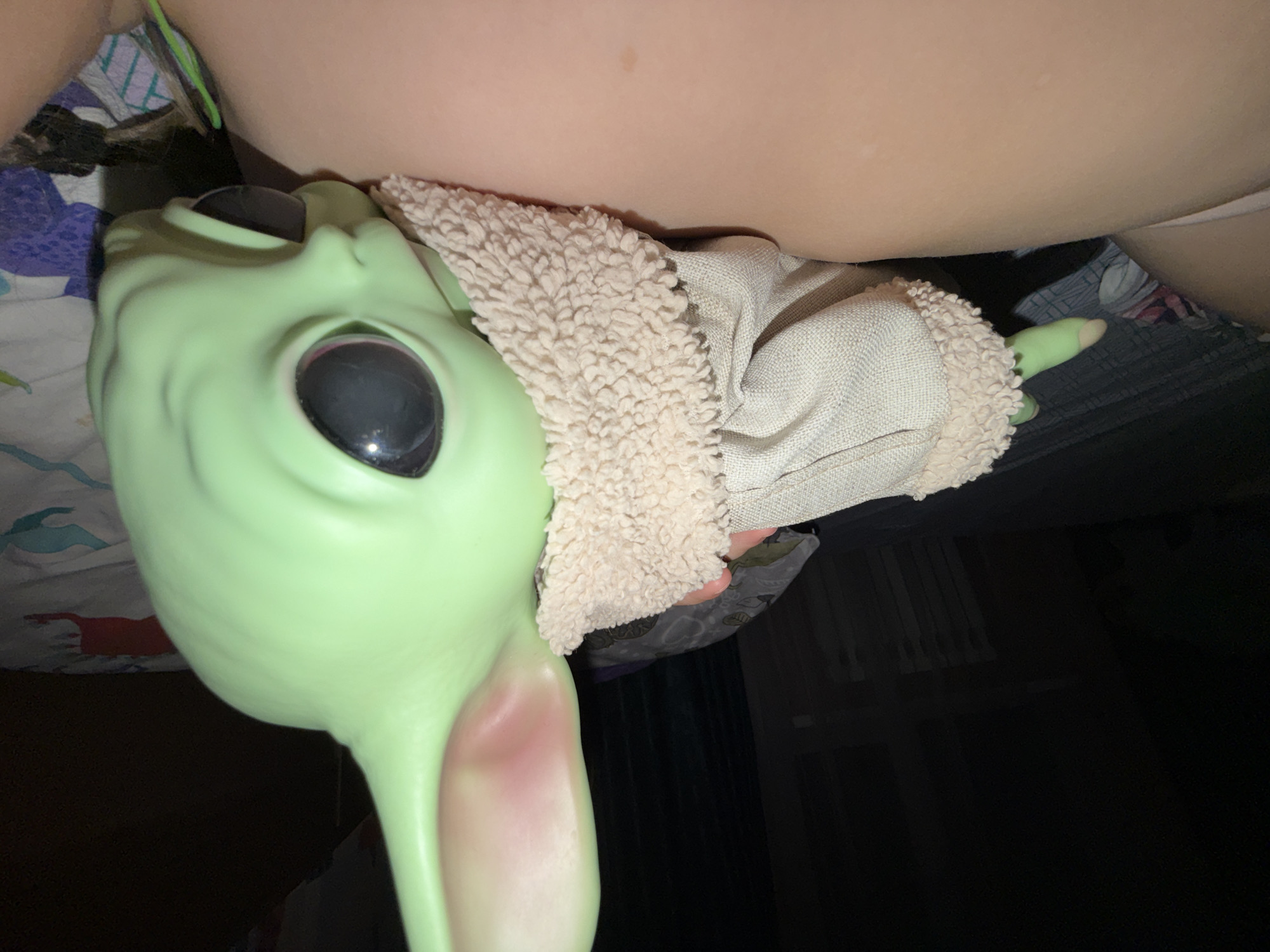 A close-up, medium-lit shot depicts a "Baby Yoda" doll, lying on a bed next to a person's thigh. The doll, dressed in a beige jacket with a fuzzy collar, has large, dark eyes and prominent ears with a reddish tint. The bed appears to have patterned sheets. It's not possible to determine the location is Chaikovsky, Russia from the image.