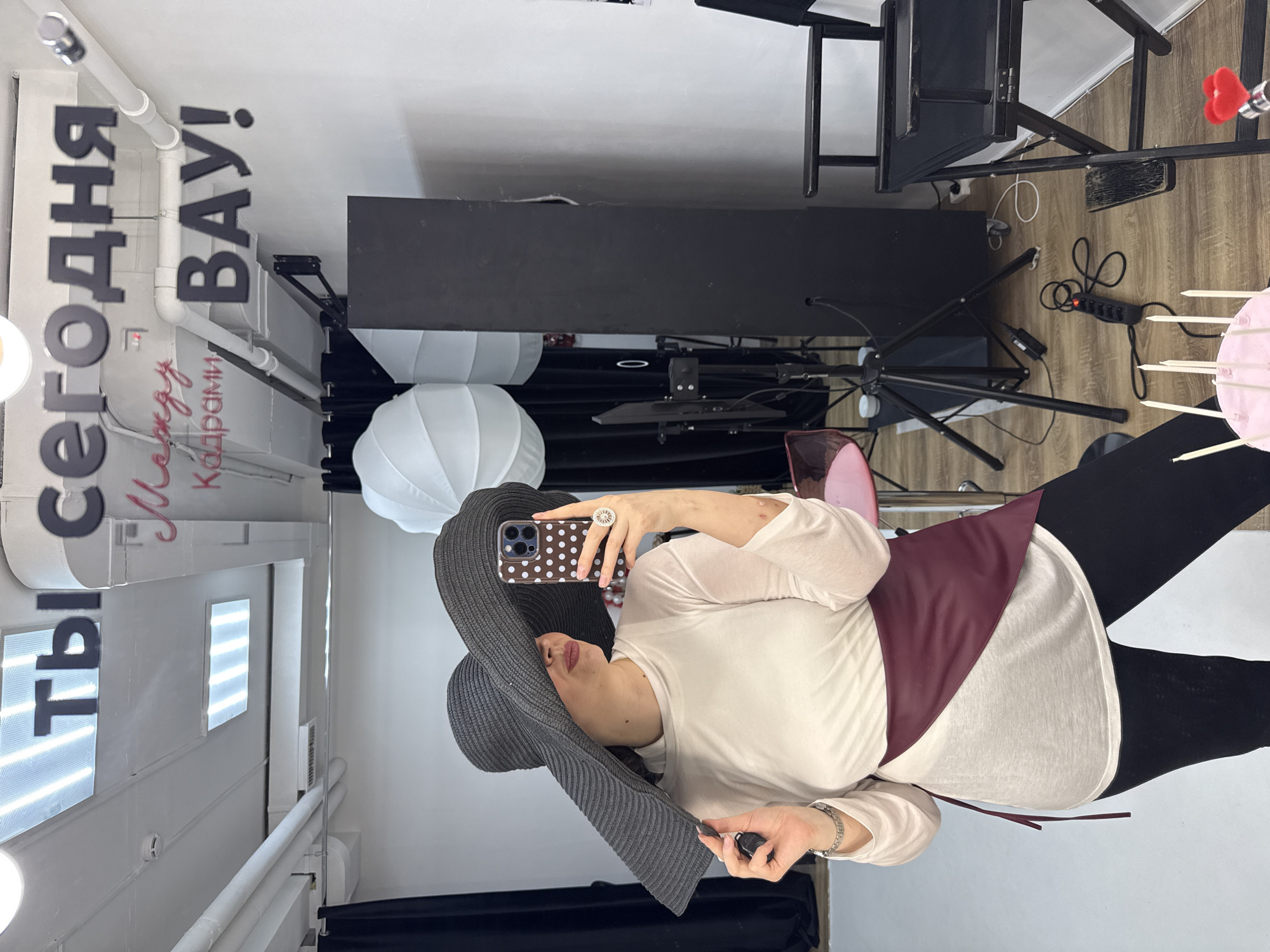 A person is taking a selfie in what appears to be a photography studio or a place with photographic equipment. They are wearing a wide-brimmed hat, a light-colored top, and black leggings or pants, and are holding a smartphone with a polka dot case. The studio setup includes lighting equipment, such as softboxes, and a director's chair. The environment indicates a professional or semi-professional photography setting. There are Cyrillic letters on the wall which can be translated as "You are today..." and then a phrase that includes the word "Frames" suggesting a slogan about capturing moments or being in the frame. Based on the Cyrillic text and other elements, this picture was taken in a location in Russia, specifically in Izhevsk, Russia.