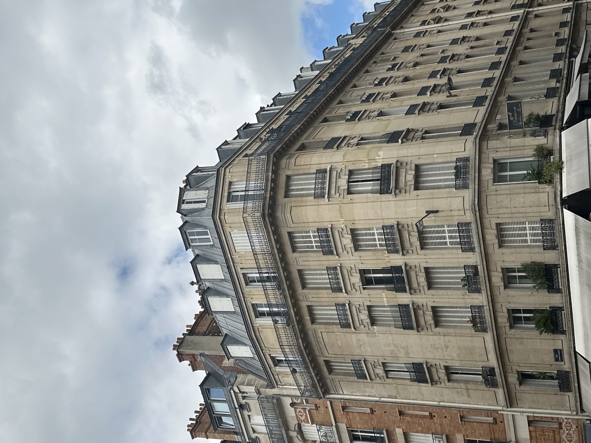 The image shows a view of the exterior of a corner building in Paris, France, with a cloudy sky overhead. The building is made of light-colored stone and features a Mansard roof with dormer windows. The facade is adorned with numerous windows, many of which have closed white shutters. Several balconies with black wrought-iron railings are visible. The building on the left is constructed with red brick. On the ground floor of the stone building, awnings are visible. A sign with ornate lettering hangs from the corner of the building, possibly indicating a business. Several potted plants are placed on window ledges and balconies. The architecture is characteristic of Parisian Haussmannian style buildings. There are no people or animals visible in the image.