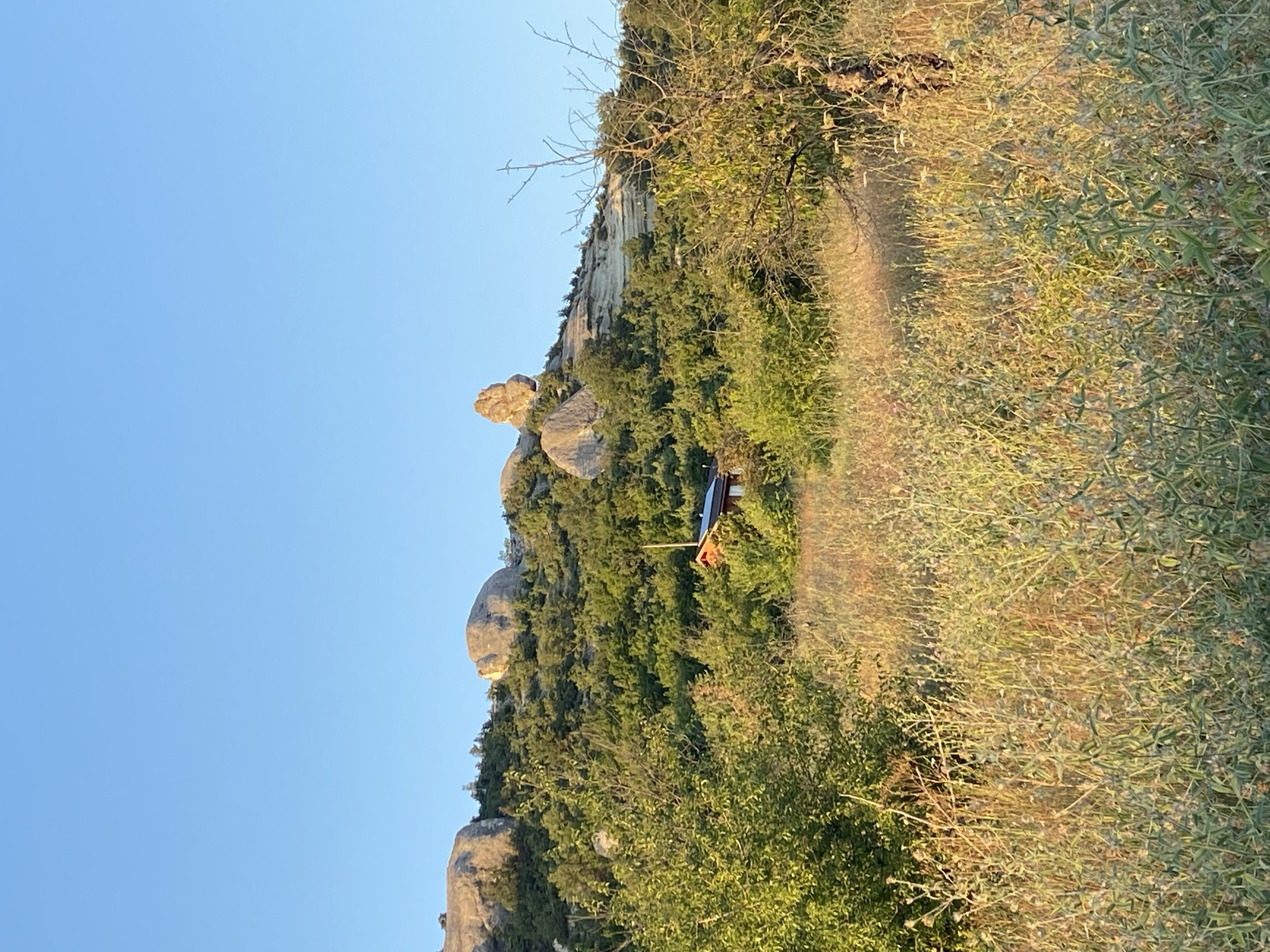 The image depicts a rural landscape bathed in the golden light of either sunrise or sunset. The foreground is dominated by a field of dry, tall grass and some green foliage, suggesting a natural, undeveloped area. In the mid-ground, a small, reddish-brown wooden structure, possibly a cabin or shed, is nestled amongst lush green trees and shrubs. Beyond the trees, a rocky, sparsely vegetated hillside rises, characterized by large, rounded boulders and layered rock formations. The sky is a clear, bright blue, indicating fair weather conditions. The overall scene conveys a sense of tranquility and natural beauty, typical of a rural or mountainous setting. There are no people visible in the image. The location is identified as being within the Krasnomakskoye rural settlement in Ukraine.