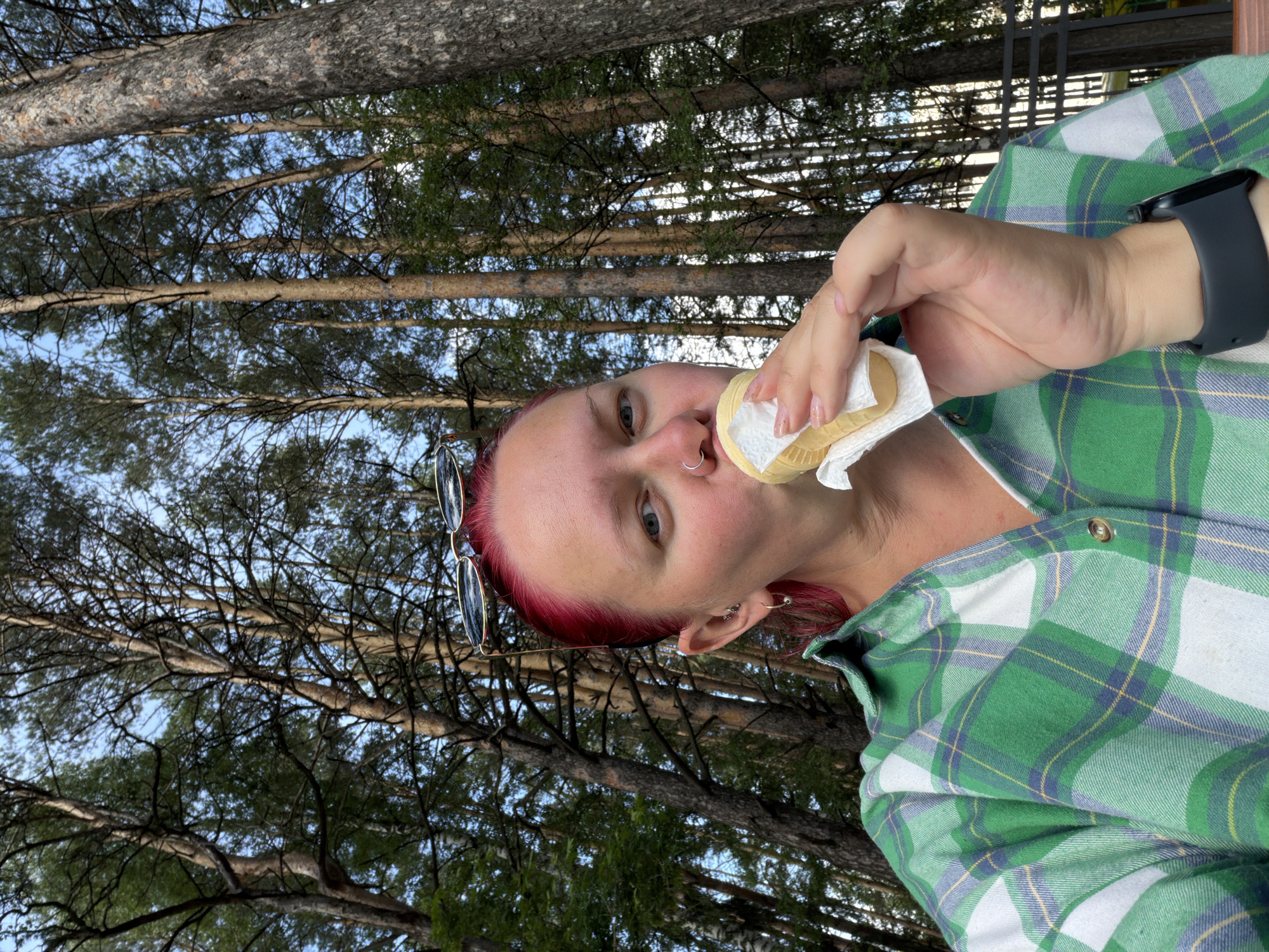 The image presents a low-angle, close-up view of an individual eating an ice cream cone in a forest setting. The person, appearing female, has short, vibrant pink or red hair, and is looking directly at the camera. Sunglasses with gold frames are perched on top of their head. They have a silver hoop nose ring and multiple earrings in their left ear. The individual wears a green, white, and blue plaid flannel shirt, and a black smartwatch is visible on their left wrist. They are holding a standard-sized ice cream cone, which is partially wrapped in a white paper napkin, and are actively biting into the ice cream.
The background consists of numerous tall, slender trees, primarily coniferous, extending upwards towards a clear, light blue sky. Sunlight is evident, illuminating the scene. In the far right background, a dark metal fence or railing is faintly visible, suggesting the proximity of a structured area within or bordering the woodland. The image itself does not contain specific visual cues or identifying landmarks to confirm its location as the city of Chaikovsky, Russia.