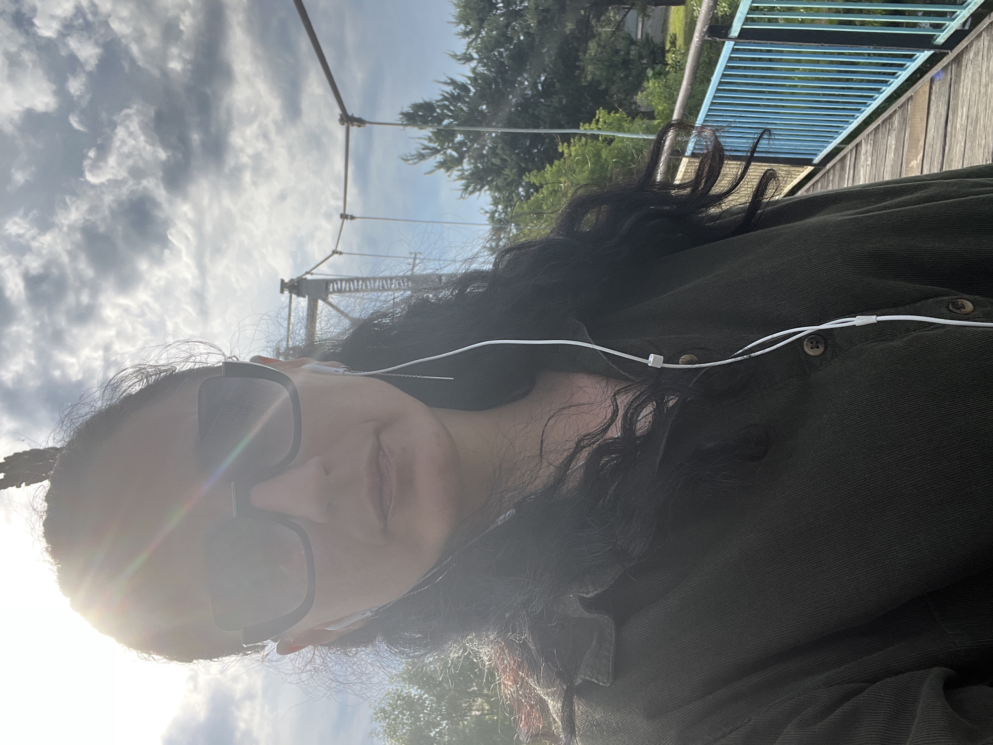 An outdoor selfie captures a person with long, dark, wavy hair, wearing a dark green, ribbed or corduroy long-sleeved shirt with prominent buttons. They are also wearing dark, square-framed sunglasses and white wired earbuds. The person is looking directly towards the camera, with a neutral or slightly smiling expression, though a prominent lens flare from the sun obscures part of their face.
The scene is set outdoors on what appears to be a bridge or elevated walkway, potentially in Kamianytsia, Ukraine. In the background, a cloudy sky with scattered white clouds is visible, illuminated by bright daylight. To the right, elements of a wooden bridge structure with distinctive blue metal railings are clearly seen, alongside lush green trees in the mid-distance. Overhead, suspension bridge cables and towers are discernible against the sky. The bright sun, positioned behind and slightly above the subject, indicates it is daytime, likely morning or late afternoon given the strong light angle. No visible text is present in the image.