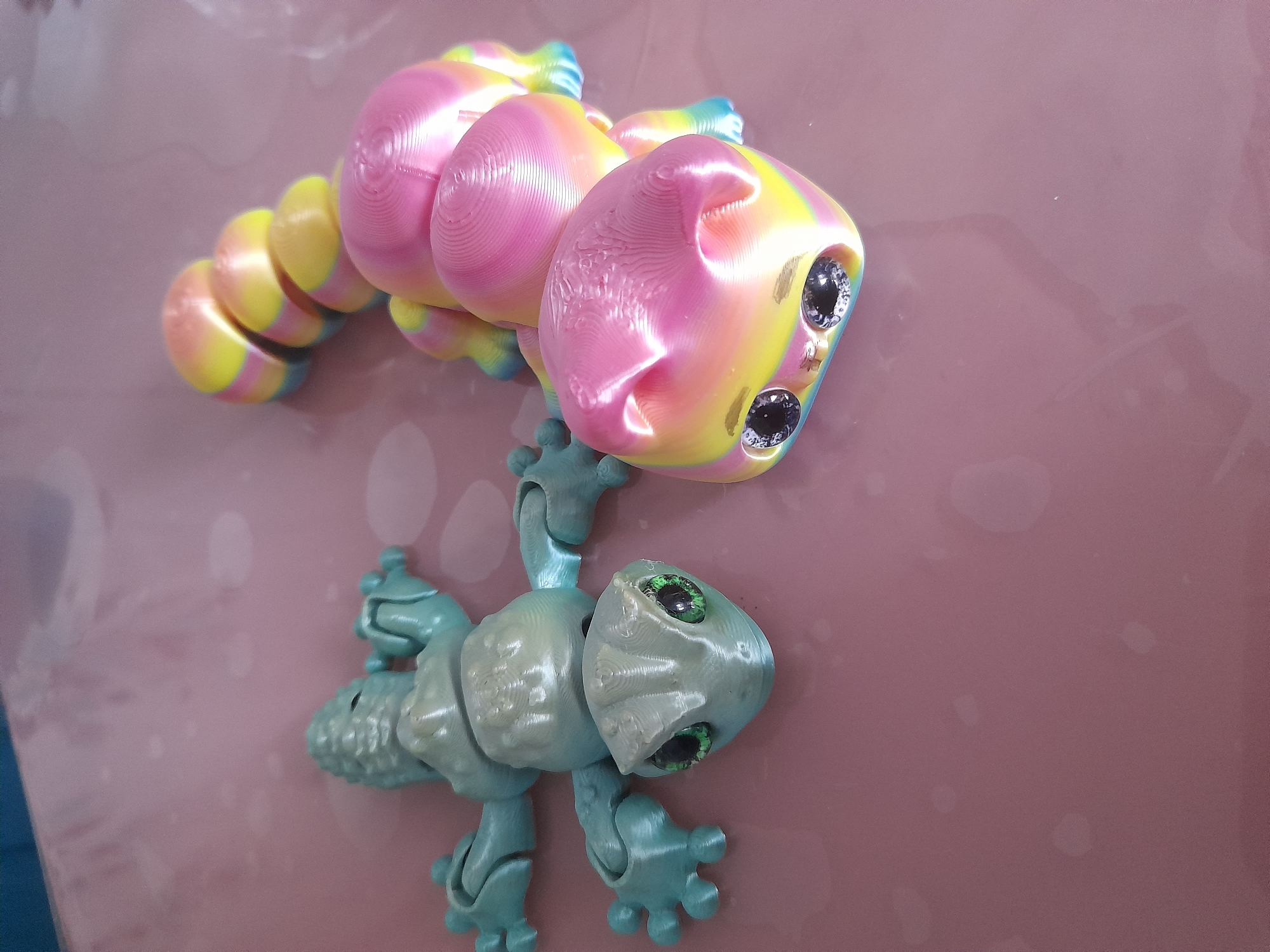 The image displays two articulated toy creatures on a pink surface. The upper creature is segmented with a smooth, rounded body, colored in yellow, pink, and blue stripes. Its head is a rounded quadrilateral with two gem-like, multifaceted black eyes.
The lower creature is a light blue-green color with a textured body. It has four limbs with three-fingered hands. Its head is also a rounded quadrilateral, with two inset green eyes. The composition shows the upper creature positioned above the lower one, with one of its limbs resting on the lower creature. The scene provides no geographical references.