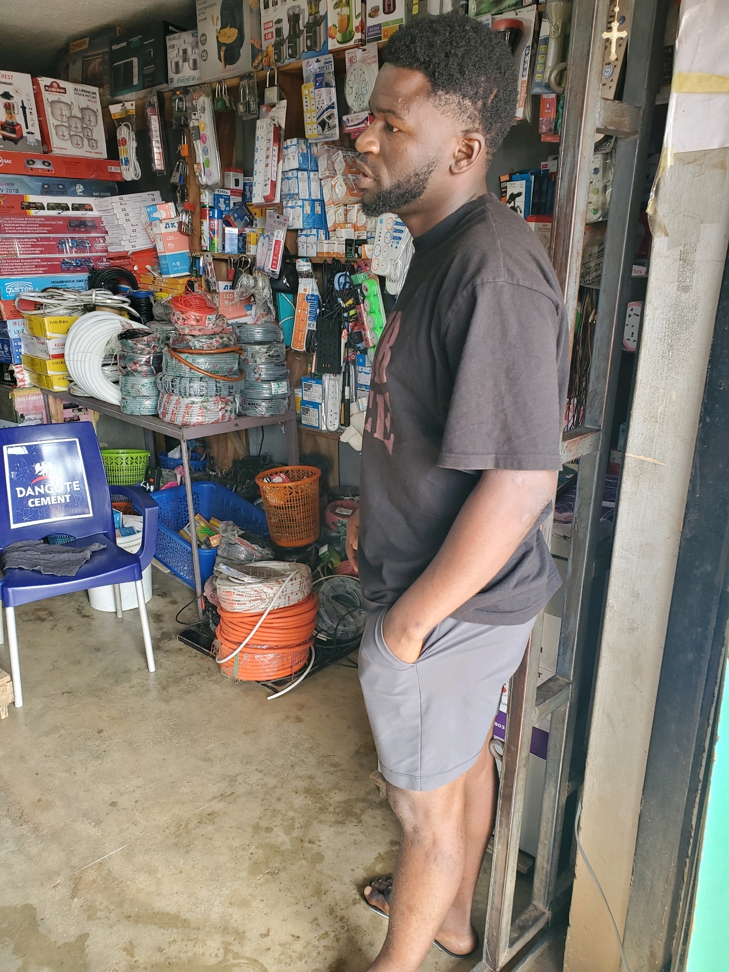 In Gura Top, Nigeria, a man stands in a retail establishment filled with various goods, likely an electronics or hardware store. The store's interior is densely packed with shelves displaying an assortment of items such as electrical cables, extension cords, electrical outlets, and what appear to be kitchen appliances. A blue plastic chair with the "DANGOTE CEMENT" logo is positioned in the foreground, and an orange basket rests on a shelf. The man, dressed in a dark t-shirt and grey shorts, has his hand in his pocket and is looking towards the right of the frame. The environment appears to be utilitarian and well-stocked, with a visible metal shelving unit and a concrete floor.