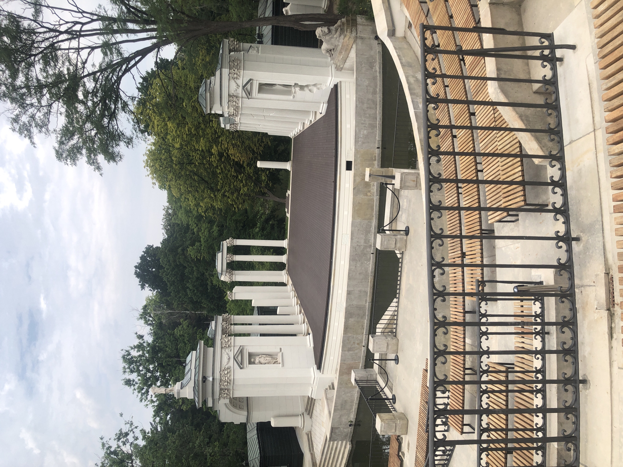 The image shows the Amphitheater in the Royal Baths Park (Łazienki Królewskie) in Warsaw, Poland. It is an outdoor amphitheater with a semi-circular seating area made of wooden benches, facing a stage area. The stage is surrounded by a moat. Behind the stage, there are classical-style white stone structures with columns, arches, and sculptures, resembling ancient ruins. The entire scene is set within a lush green park with mature trees. The sky is overcast. There are no people visible in the image.