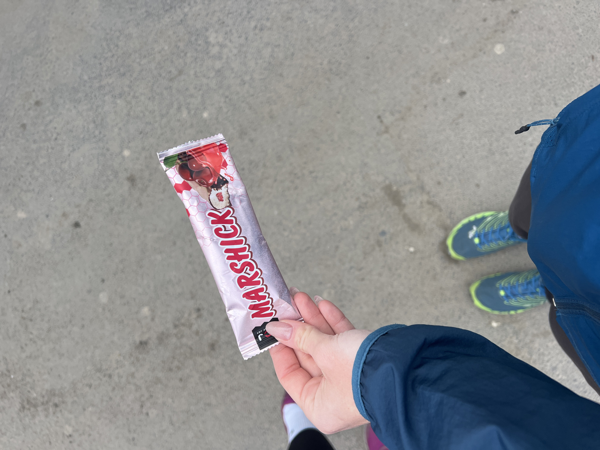 A person, likely an adult female, is holding a cherry-flavored "Marshick" brand ice cream bar in its original packaging. The person is wearing blue athletic gear and blue running shoes with neon green accents. The person is standing on a gray concrete surface, potentially a sidewalk, on the streets of the city of Perm, Russia, based on the Russian language on the package.