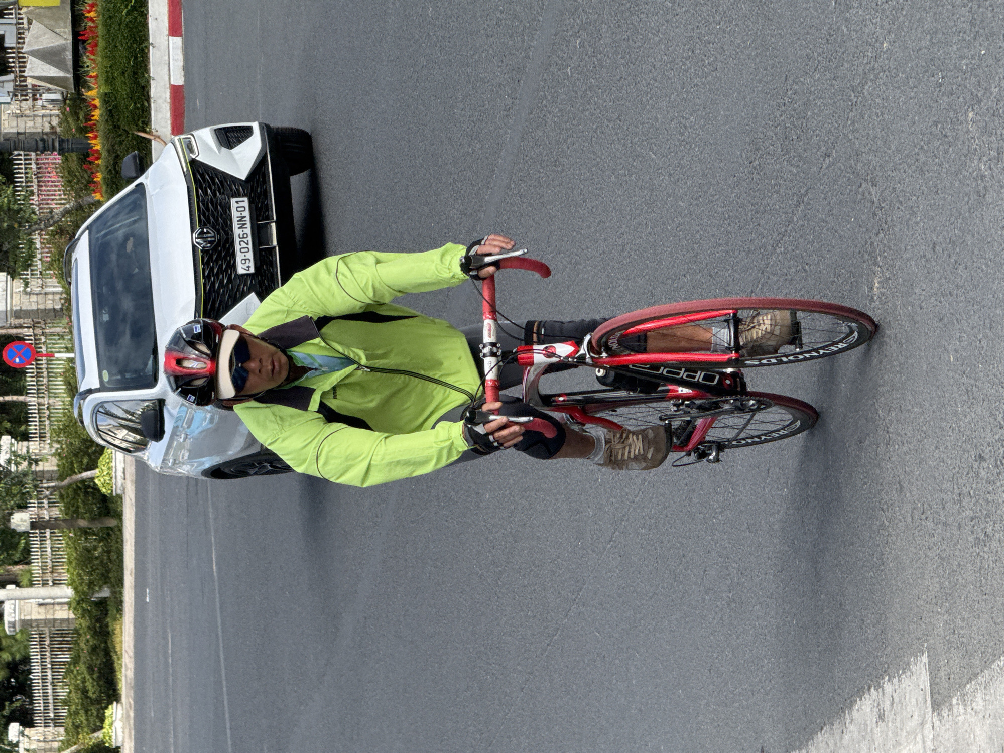 A man wearing a neon green jacket, sunglasses, and a red and black helmet rides a red bicycle. The man's hands are gloved and he is holding the handlebars. His feet are in brown cycling shoes. He is riding on an asphalt road with a white line on the left side of the image. In the background, a white car with license plate 49-026-NN-01 is stopped on the road. The car is facing forward and its headlights are on. Behind the car, there is a white fence and some green bushes. The image appears to have been taken in Da Lat, Vietnam, as indicated by the license plate format and the mountainous terrain visible in the distance.