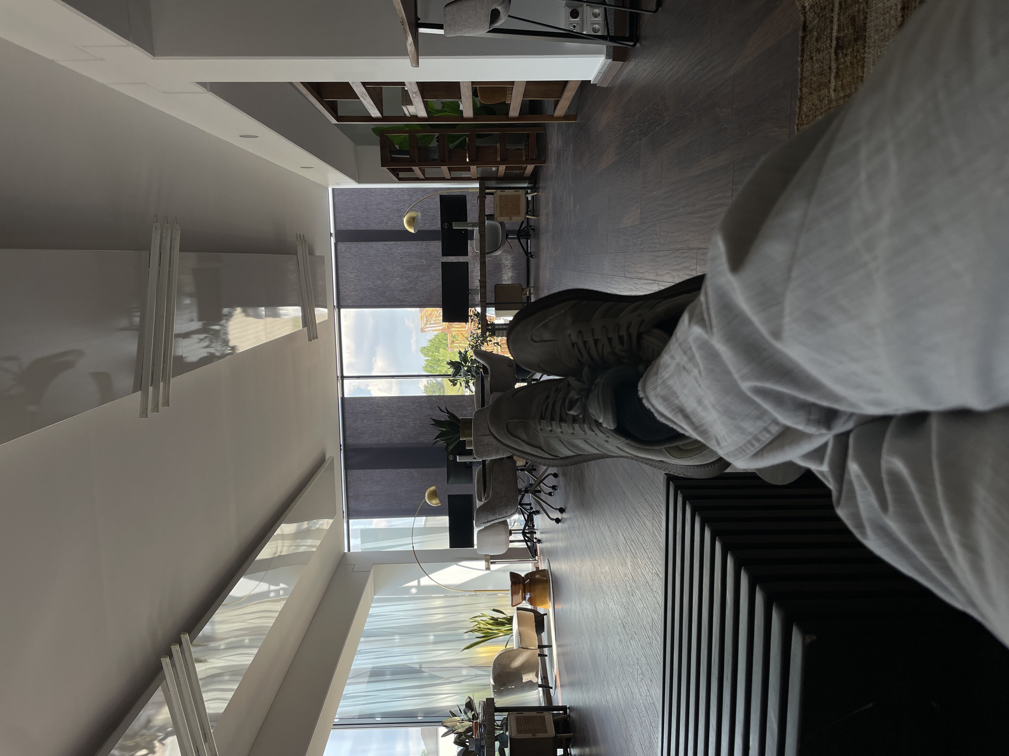 A person is seated in an office space, with their feet and legs visible in the foreground. The office features a large window providing natural light, with multiple workstations visible in the background. The window shows a cityscape with trees and buildings. The floor is dark hardwood, and there are architectural elements such as bookshelves and modern lighting fixtures.