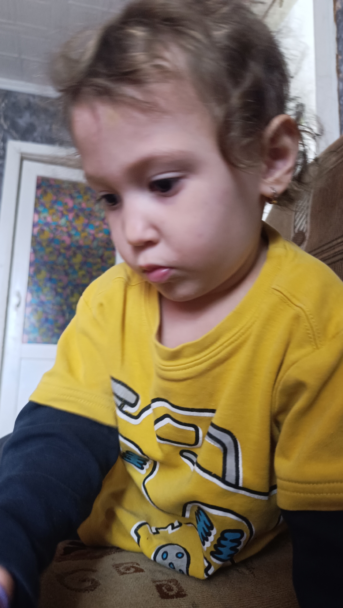 The image is a close-up portrait of a young child, likely a toddler, with light brown hair. The child is wearing a yellow long-sleeved shirt with a cartoon design. The shirt is predominantly yellow with black sleeves and features a graphic with white and blue elements. The child's face is turned to the side and downwards, with a slightly pursed mouth. The background is blurred, but appears to be an indoor setting with some patterned wallpaper and a doorway with colored glass. The child is seated on a patterned fabric surface, possibly a sofa or chair. There is no discernible indication of the city of Karsha, Russia, in the image.