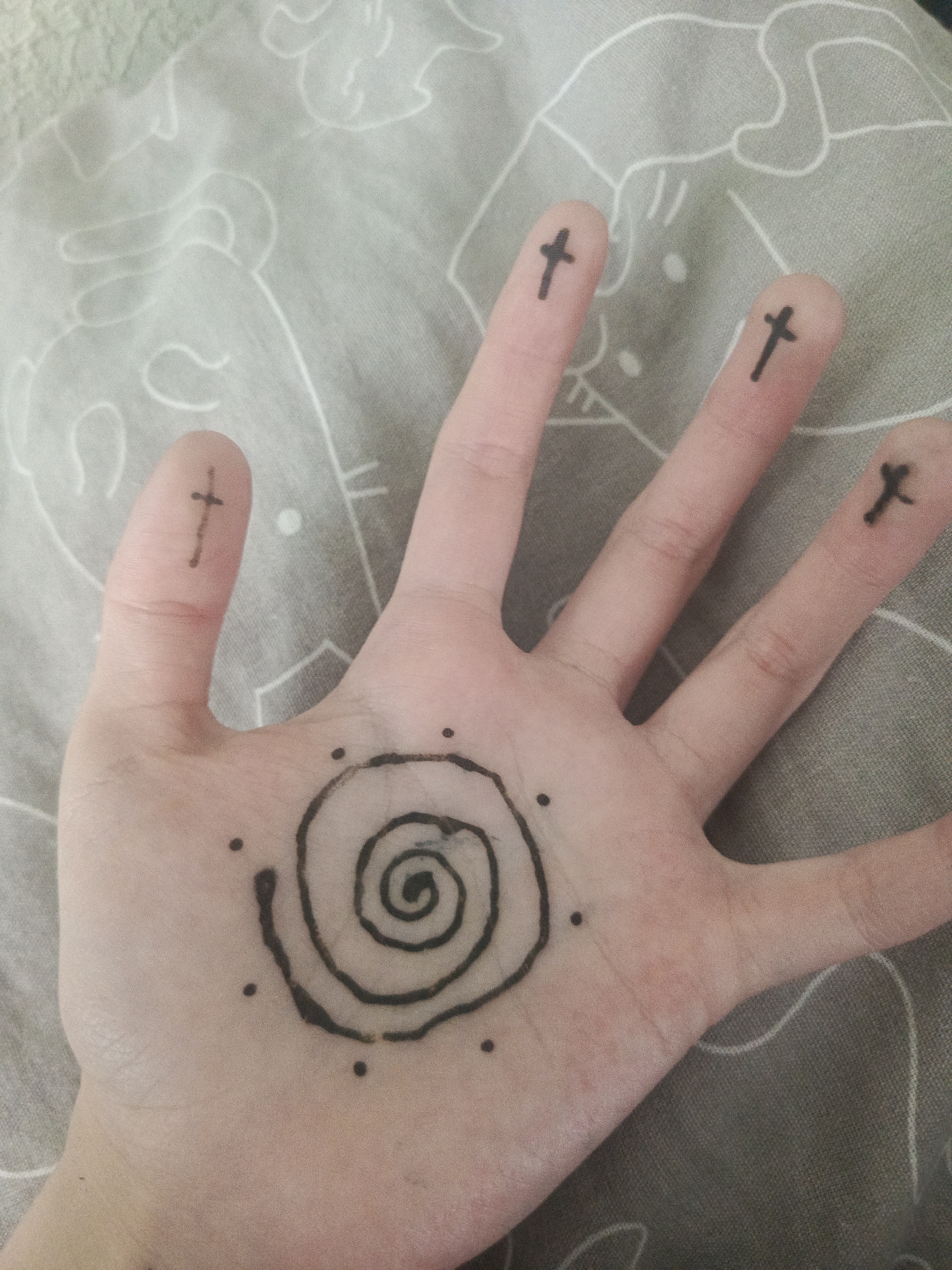 An open human palm is displayed against a patterned grey fabric. On the palm is a black spiral design encircled by six black dots. Each fingertip is marked with a small black cross. The location is Unknown, Unknown.