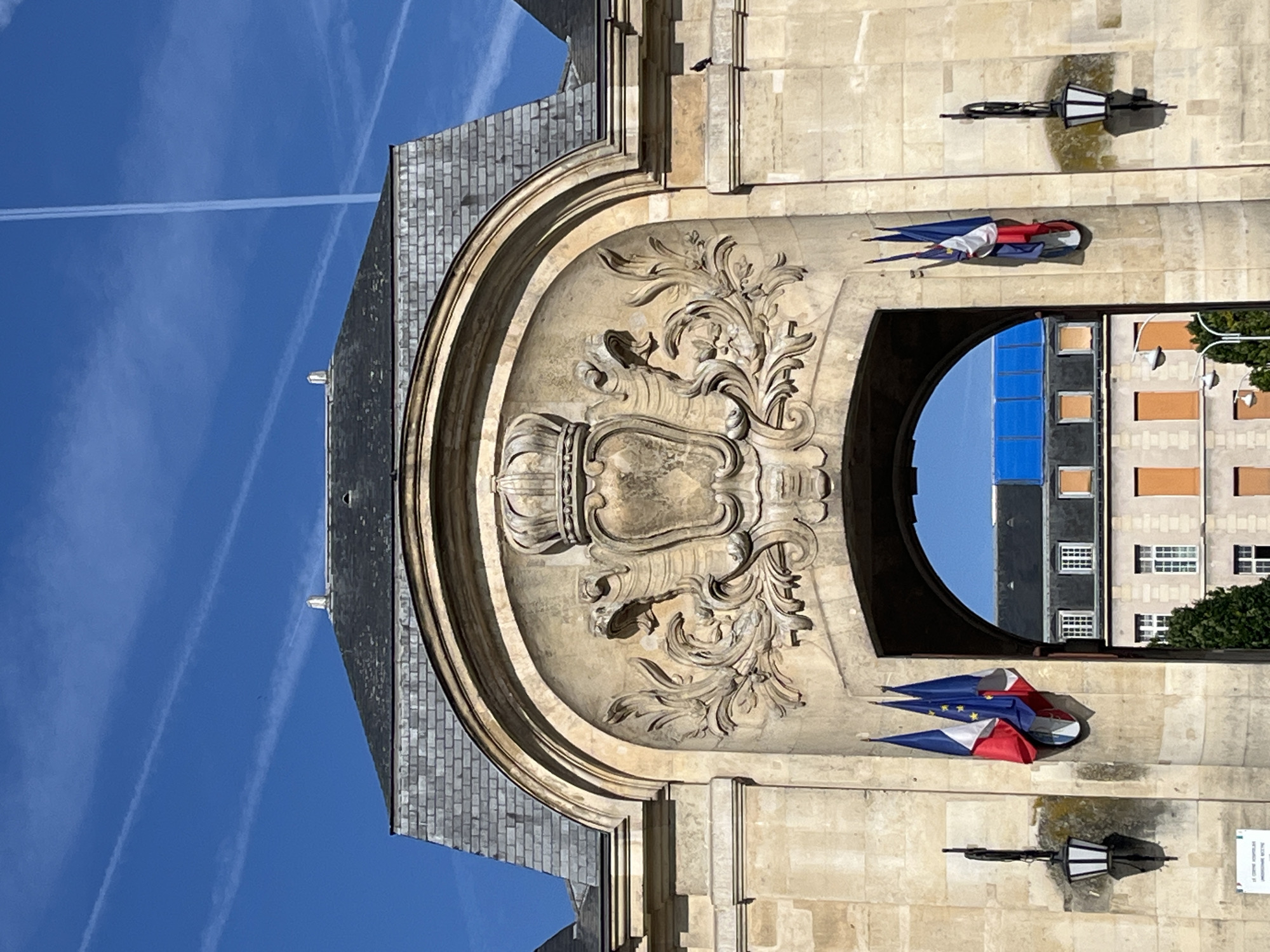 The image shows the facade of a building with a large arched entrance. Above the arch, there is a stone relief featuring a crown and ornate scrollwork, flanked by two winged figures. On either side of the arch, there are black lampposts. Two French flags are hanging on the wall to the left of the arch and one flag is hanging to the right of the arch. Through the arch, a street with buildings and trees can be seen. The sky is blue with white contrails. The architectural style suggests it is a historical building, possibly a government building or a monument. The flags indicate a French location. The building is made of light-colored stone. The overall impression is one of grandeur and historical significance. This is the Hôtel de Ville (City Hall) of Le Kremlin-Bicêtre, France.