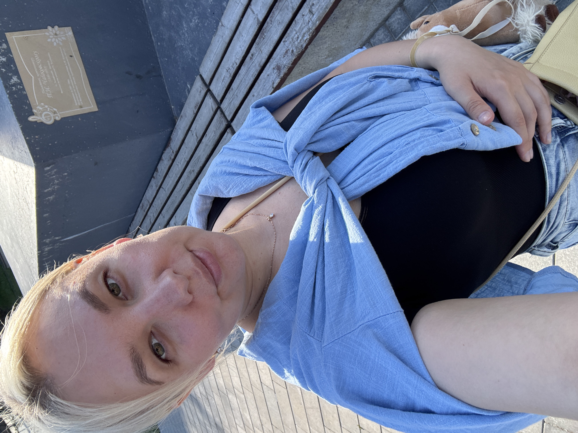 A young woman with blonde hair and green eyes is taking a selfie outdoors. She is wearing a light blue unbuttoned shirt tied at her waist over a black tank top and denim shorts. A delicate necklace with a small diamond pendant is visible. To her left, a plaque is mounted on a dark blue surface. The plaque is light brown with some text and an engraved image of a flower and a treble clef. The text on the plaque appears to be in Cyrillic script and reads: "П.И. Чайковскому" (P.I. Tchaikovsky) followed by dates and additional inscription. She is seated on wooden planks which appear to be part of a bench or walkway. A small stuffed toy and a tan purse are visible behind her right arm. The lighting suggests it is daytime, possibly late afternoon given the warm sunlight. The overall mood seems relaxed and casual.