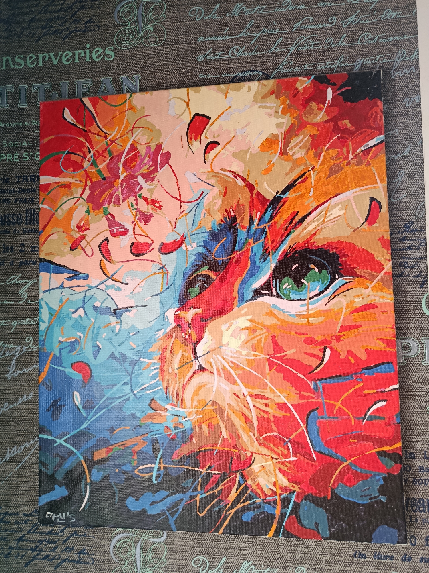 The image displays a colorful, abstract painting of a cat's face, predominantly featuring vibrant shades of red, orange, yellow, blue, and green. The cat's eye is a prominent focal point, rendered in striking green with hints of blue. The painting is positioned on a dark gray textured surface that appears to be a fabric with faint, light-blue cursive writing and decorative elements. This surface serves as a backdrop, partially obscuring text on the left side of the image, including the word "nserviries" and "TIT IFAN". The painting is likely an acrylic or oil on canvas, given its texture and the visible brushstrokes. There is no indication or contextual information within the image that connects it to the city of Apatity, Russia, or any specific urban environment. The scene is static and focuses solely on the artwork and its immediate presentation.